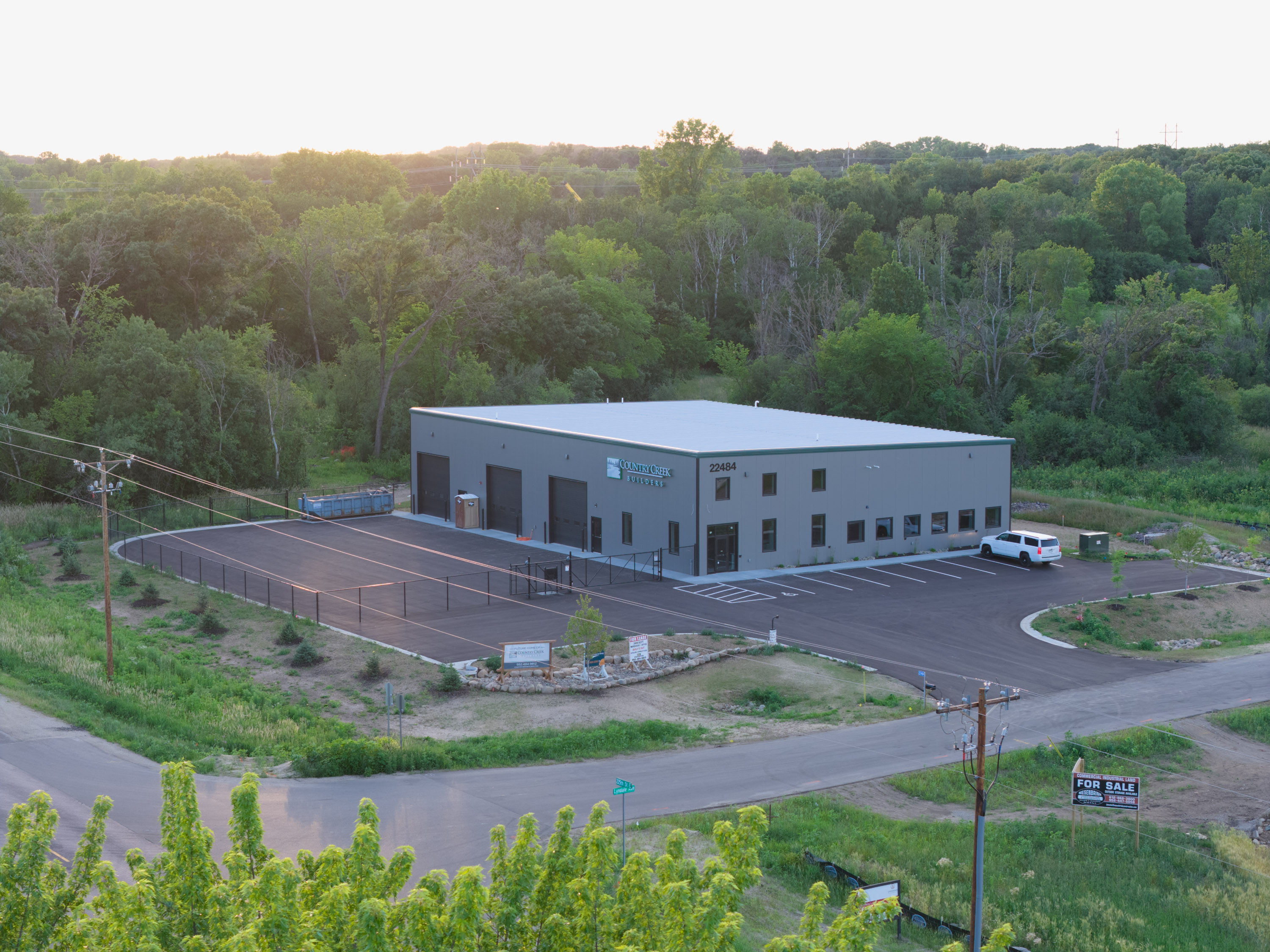 Country Creek Builders new Lakeville MN showroom building exterior, aerial view at dusk showing completed gray metal panel construction with green trim, parking lot, and wooded surroundings