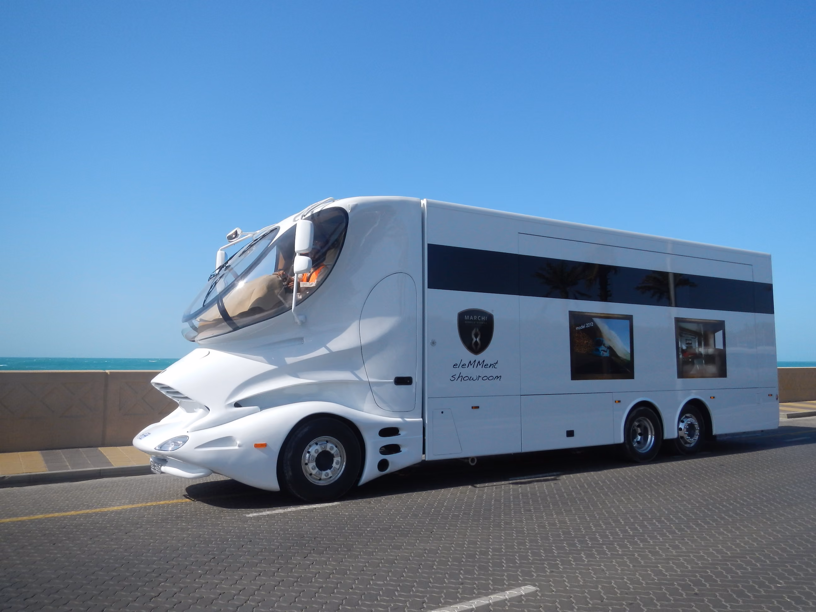 White Luigi Colani inspired design truck parked by ocean under blue sky.