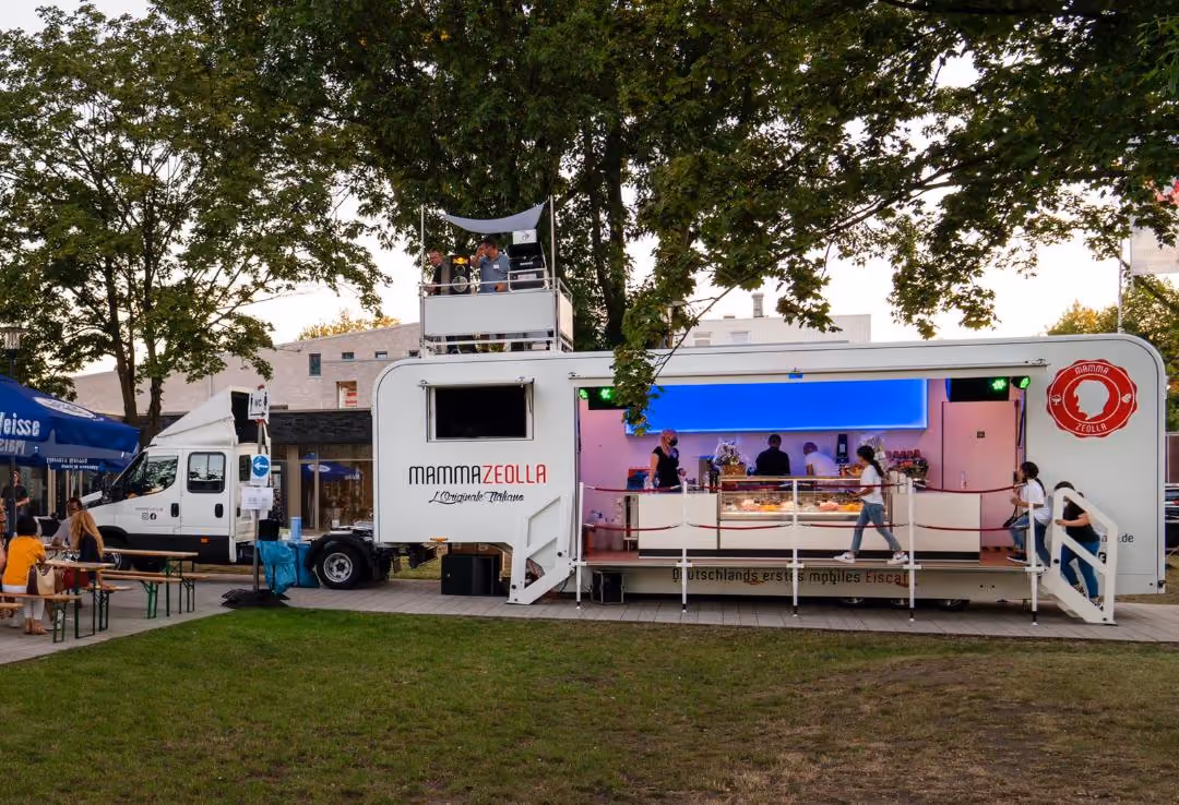 Moderner Eis-Truck "Mamma Zeolla" mit rosa Innenbeleuchtung und Besucher unter grünen Bäumen.