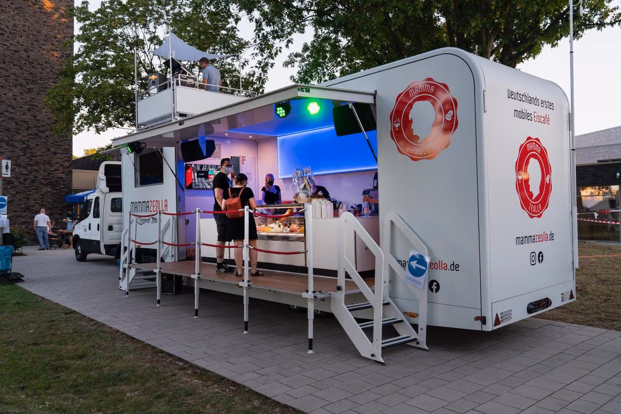 Moderner Eis-Truck "Mamma Zeolla" mit rosa Innenbeleuchtung und Besucher unter grünen Bäumen.