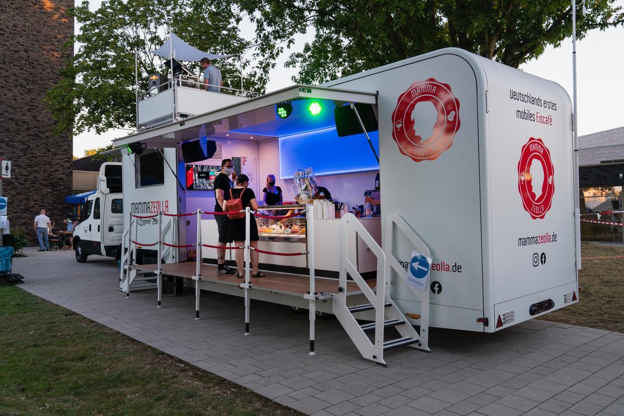 Moderner Eis-Truck "Mamma Zeolla" mit rosa Innenbeleuchtung und Besucher unter grünen Bäumen.