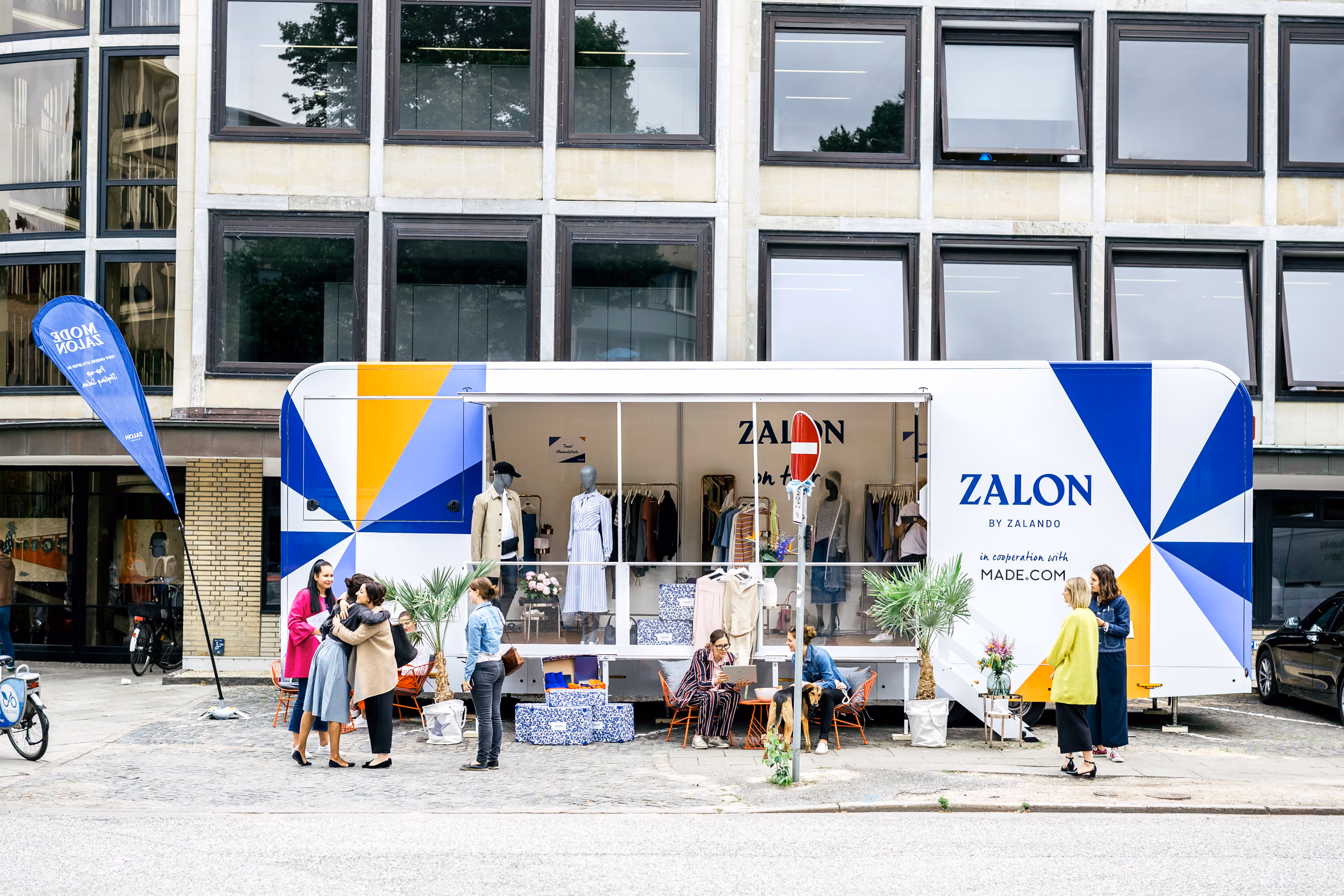 Ein Pop-Up-Store-PKW-Anhänger von Zalando mit blau-gelbem Design auf einer urbanen Straße, Menschen besuchen den Pop-up-Shop.