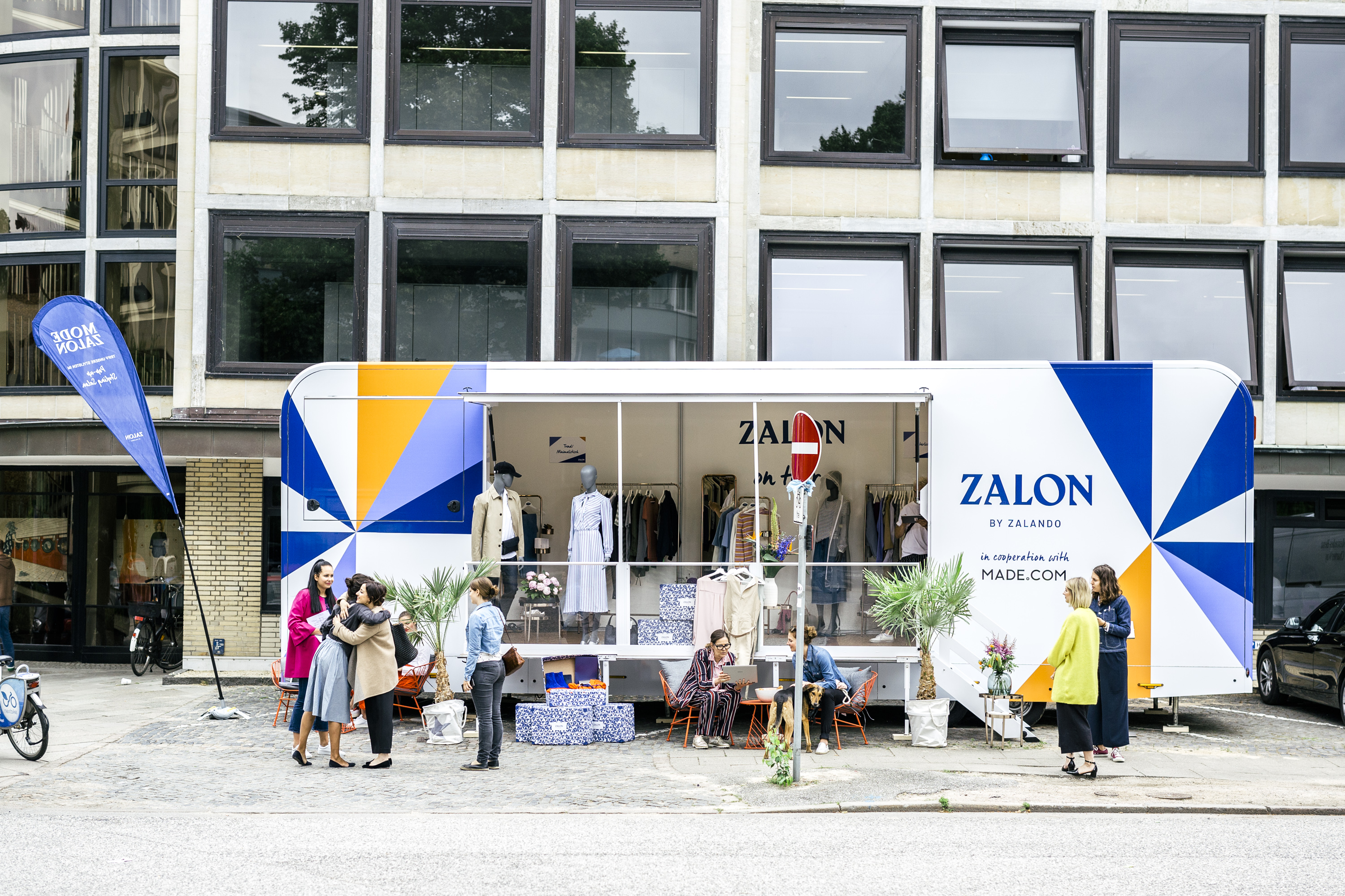 Ein Pop-Up-Store-PKW-Anhänger von Zalando mit blau-gelbem Design auf einer urbanen Straße, Menschen besuchen den Pop-up-Shop.
