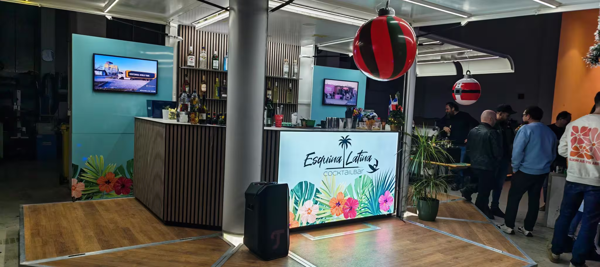 Moderne Cocktailbar in Cube-PKW-Anhänger mit tropischem Design, Gästen und hängenden roten Dekorationskugeln.