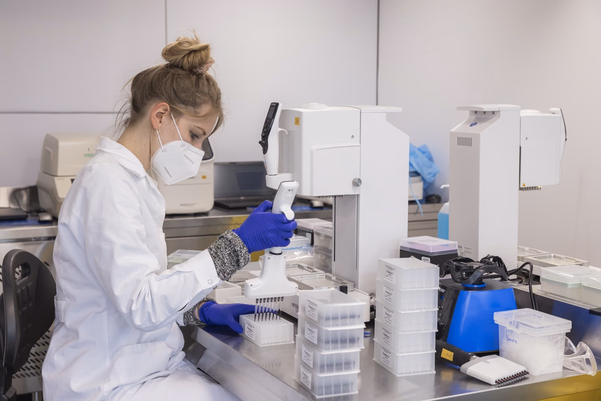 Lab technician in white coat and blue gloves working with samples and medical equipment.
