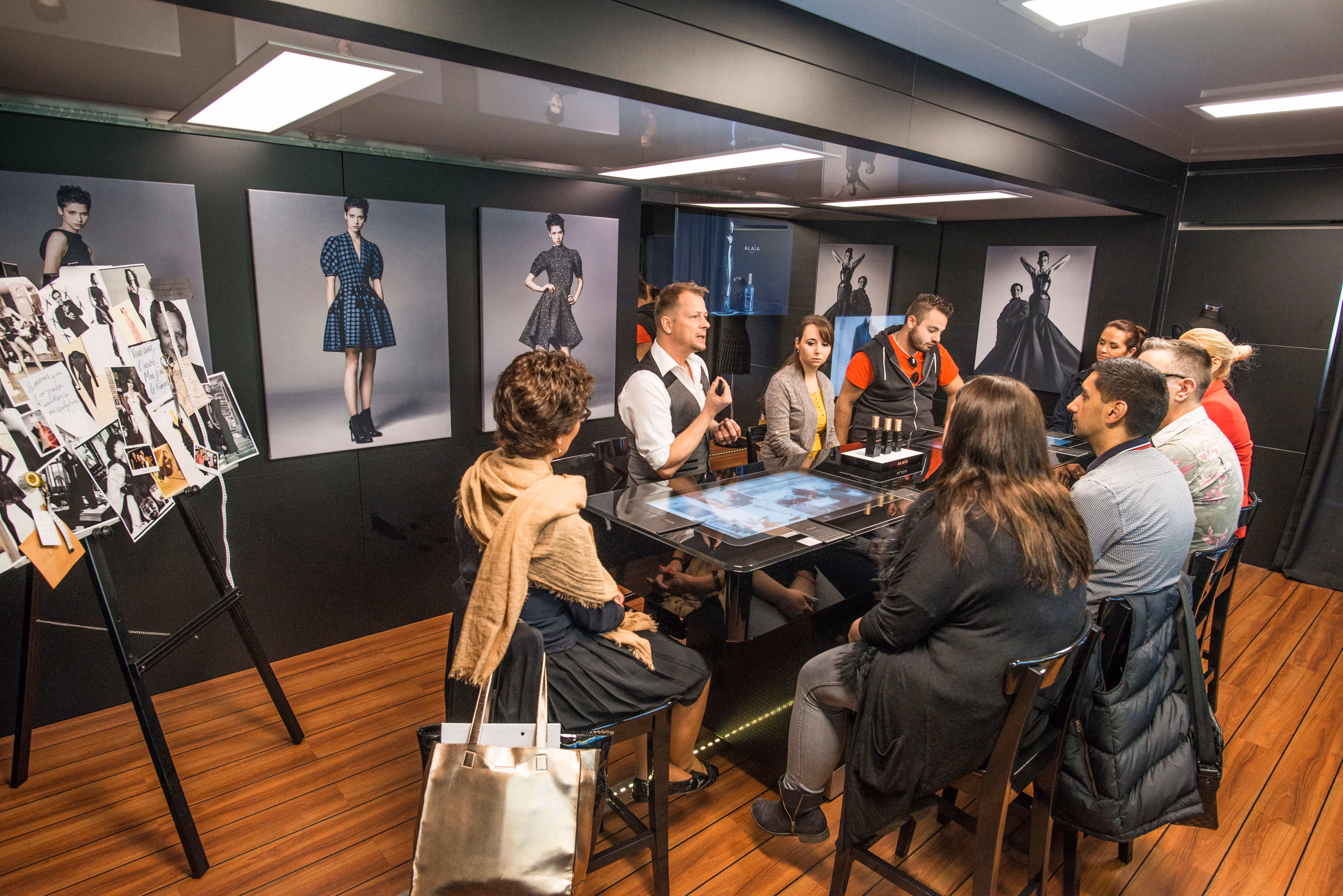 A man presents a perfume collection to representatives in a modern design studio, with sketches and images in the background.