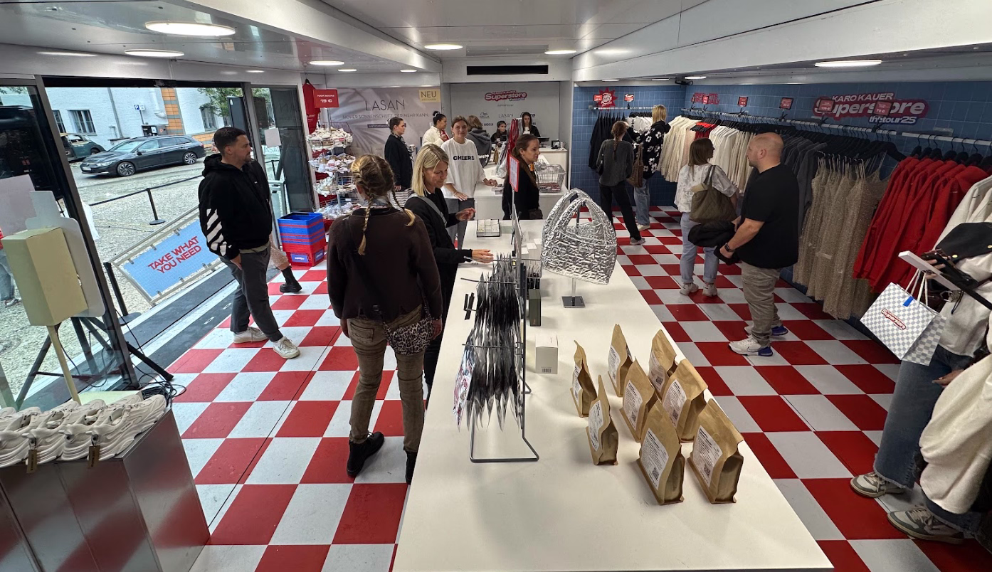 Moderner Laden in Sattelauflieger mit Bezahlzone im Hintergrund, Kundinnen shoppen Kleidung und Accessoires an Verkaufstischen.