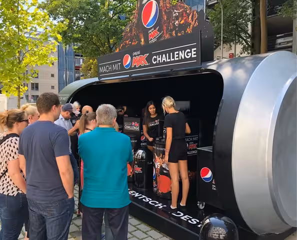 Promoter stehen in PKW-Anhänger in Form einer Pepsi-Dose und verteilen Pepsi-Dosen an Passanten.