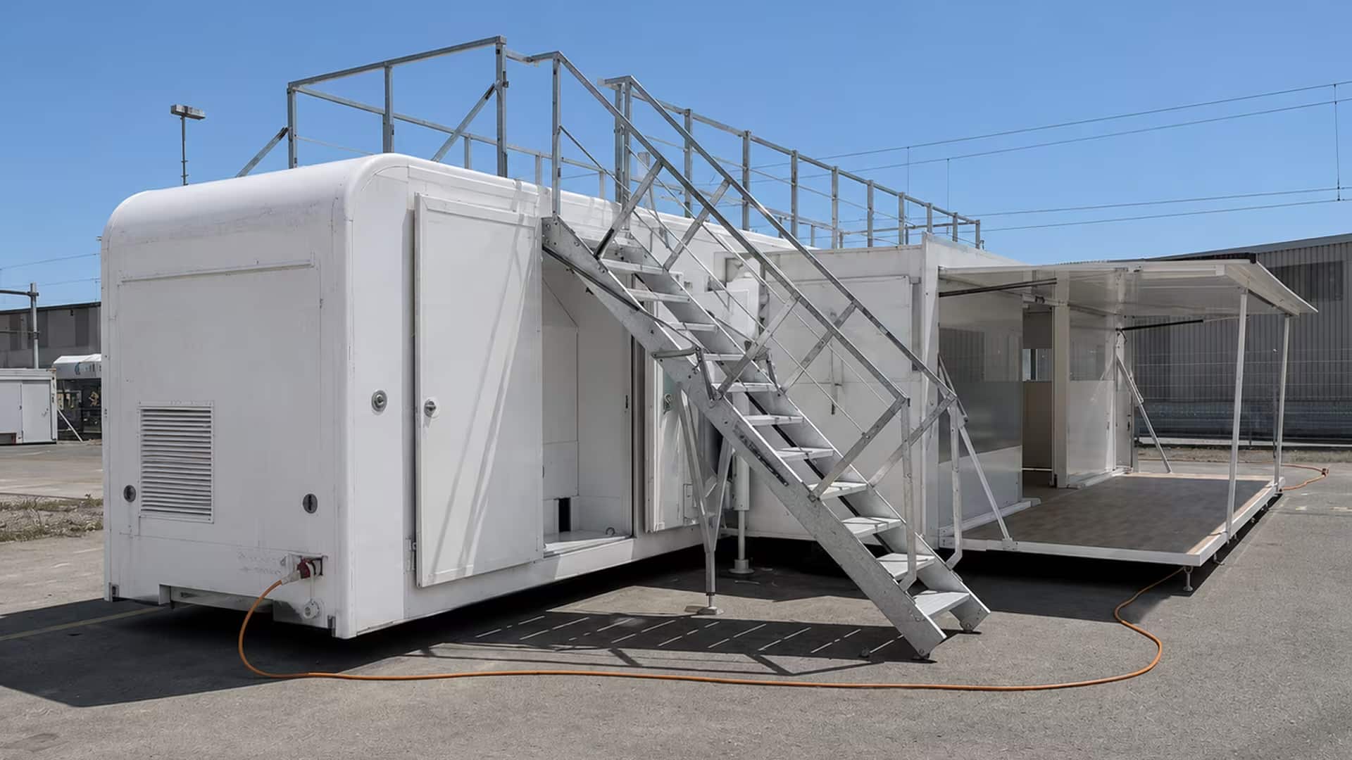 Weißer Marchi Container ohne Branding mit seitlicher Öffnung. Außen führt eine Metalltreppe auf eine Dachterrasse mit Geländer.