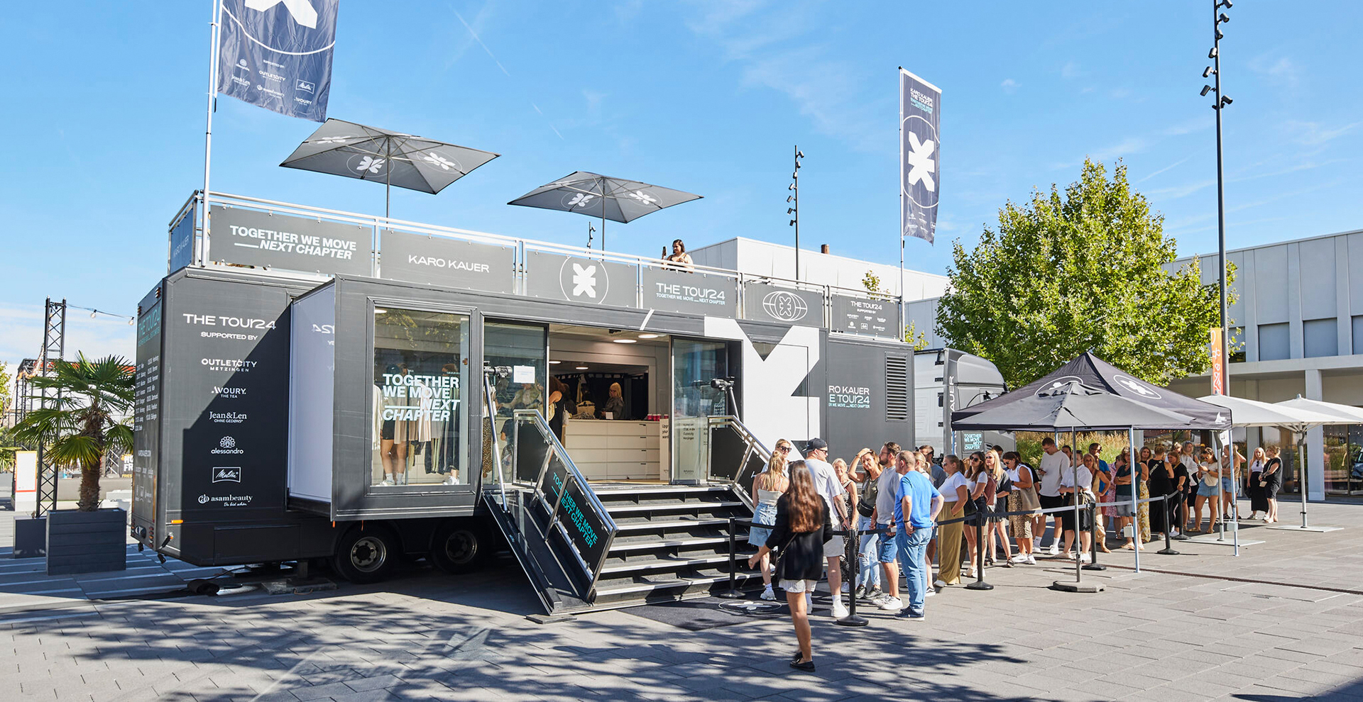 Moderner schwarzer Popup-Store-Truck von Karo Kauer mit Markenlogos und Menschenmenge an sonnigem Platz.