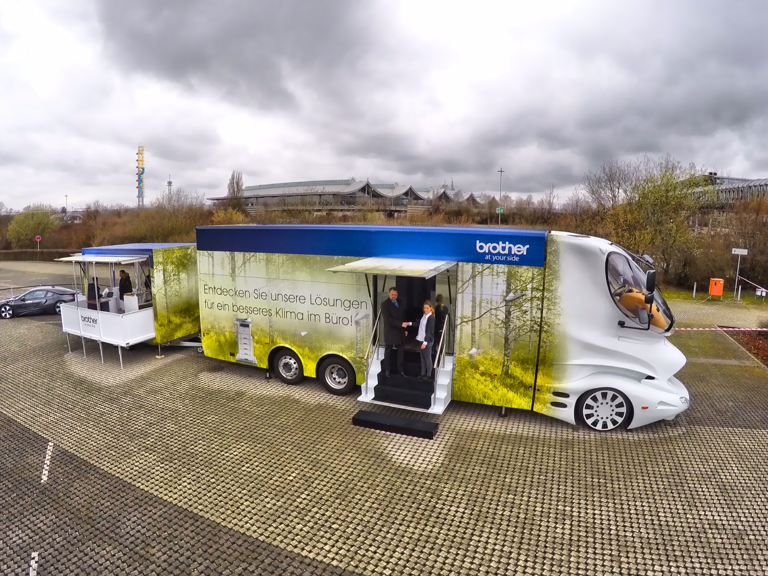 Der eleMMent Visione Designtruck von brother steht mit geöffneter Tür auf einem Parkplatz. Auf der Seite des Showtrucks steht "Entdecken Sie unsere Lösungen für ein besseres Klima im Büro!"