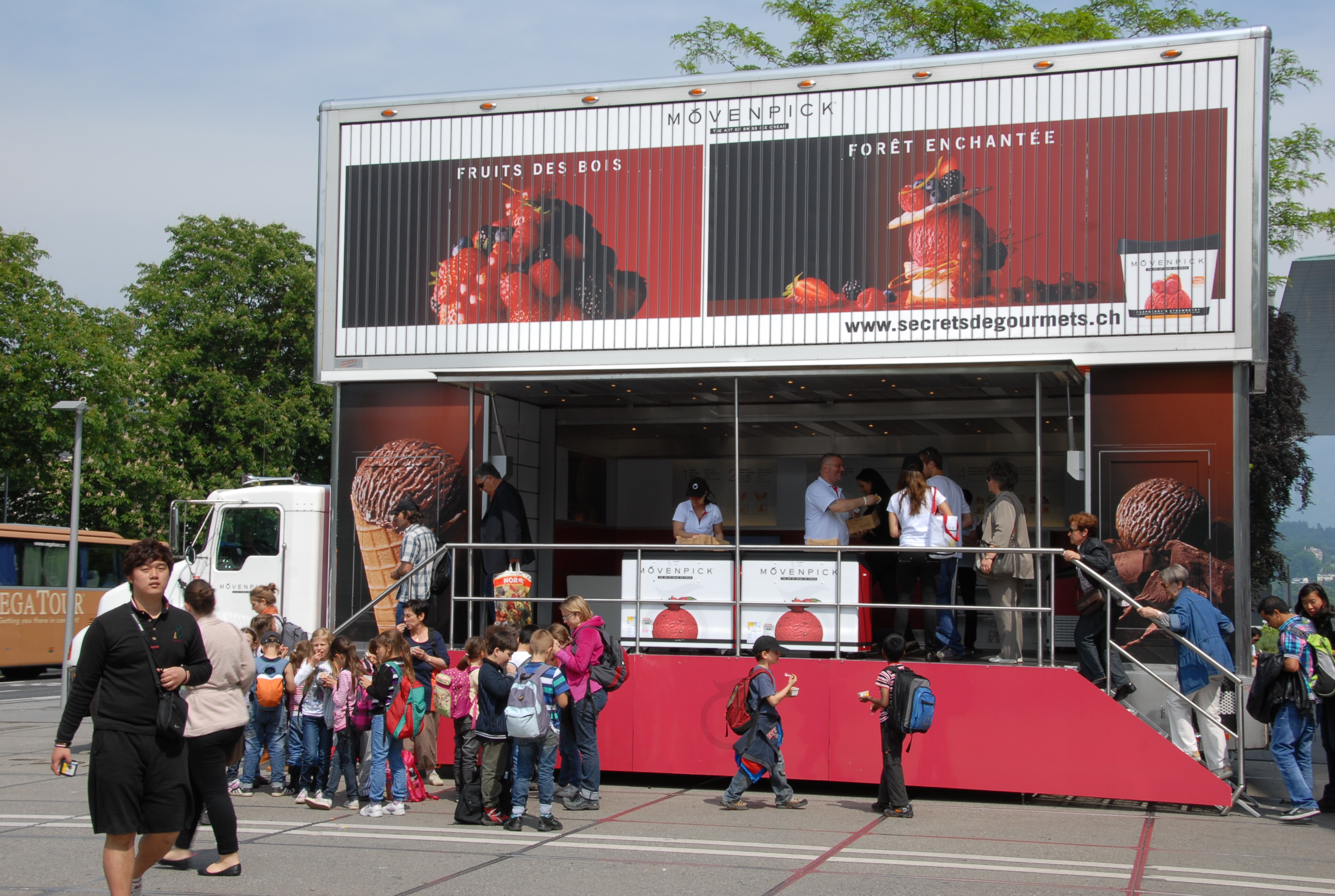 Zweistöckiger Eistruck von Mövenpick mit Waldfrucht-Branding und einer Schlange anstehender Menschen.