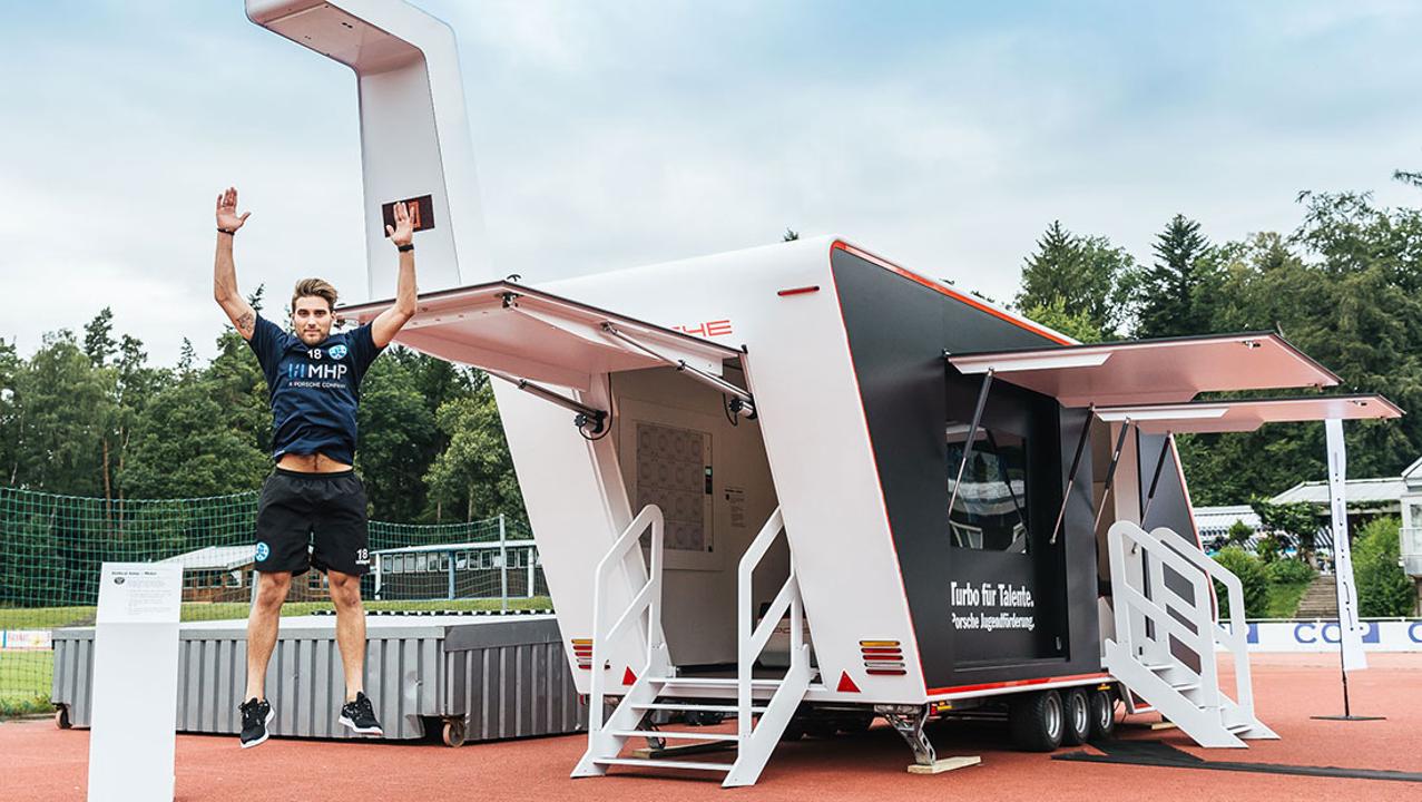 Ein geöffneter, schwarz-weißer Promotion Anhänger steht auf einem Sportplatz. Ein Athlet springt davor jubelnd in die Höhe und hält ein Tablet in der Hand.