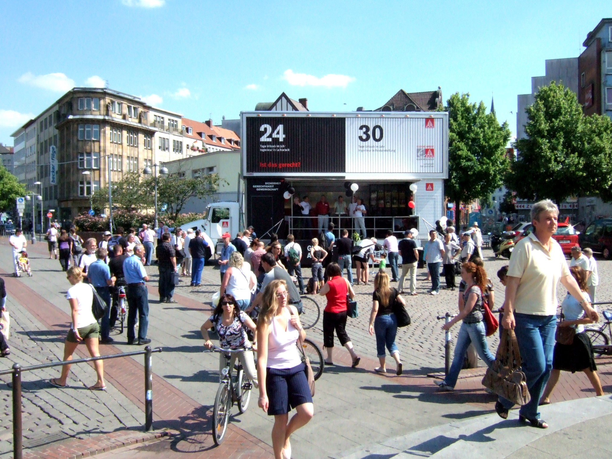 Belebter Marktplatz mit vielen Passanten an einem sonnigen Tag. Im Hintergrund steht ein großer Showtruck mit zweistöckigem Sattelauflieger.