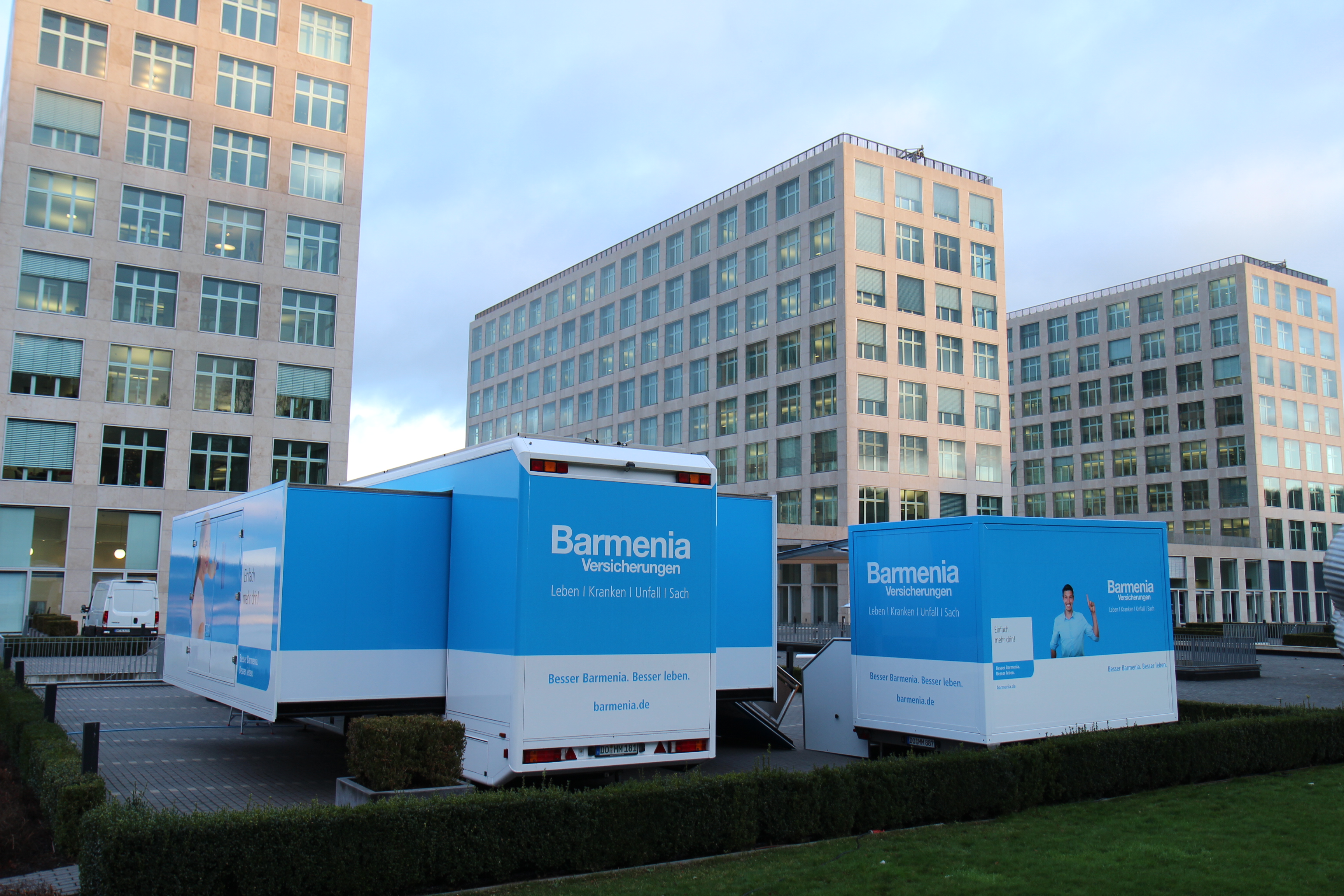 Zwei blaue Barmenia-Showtruck-Fahrzeuge vor modernen Bürogebäuden.