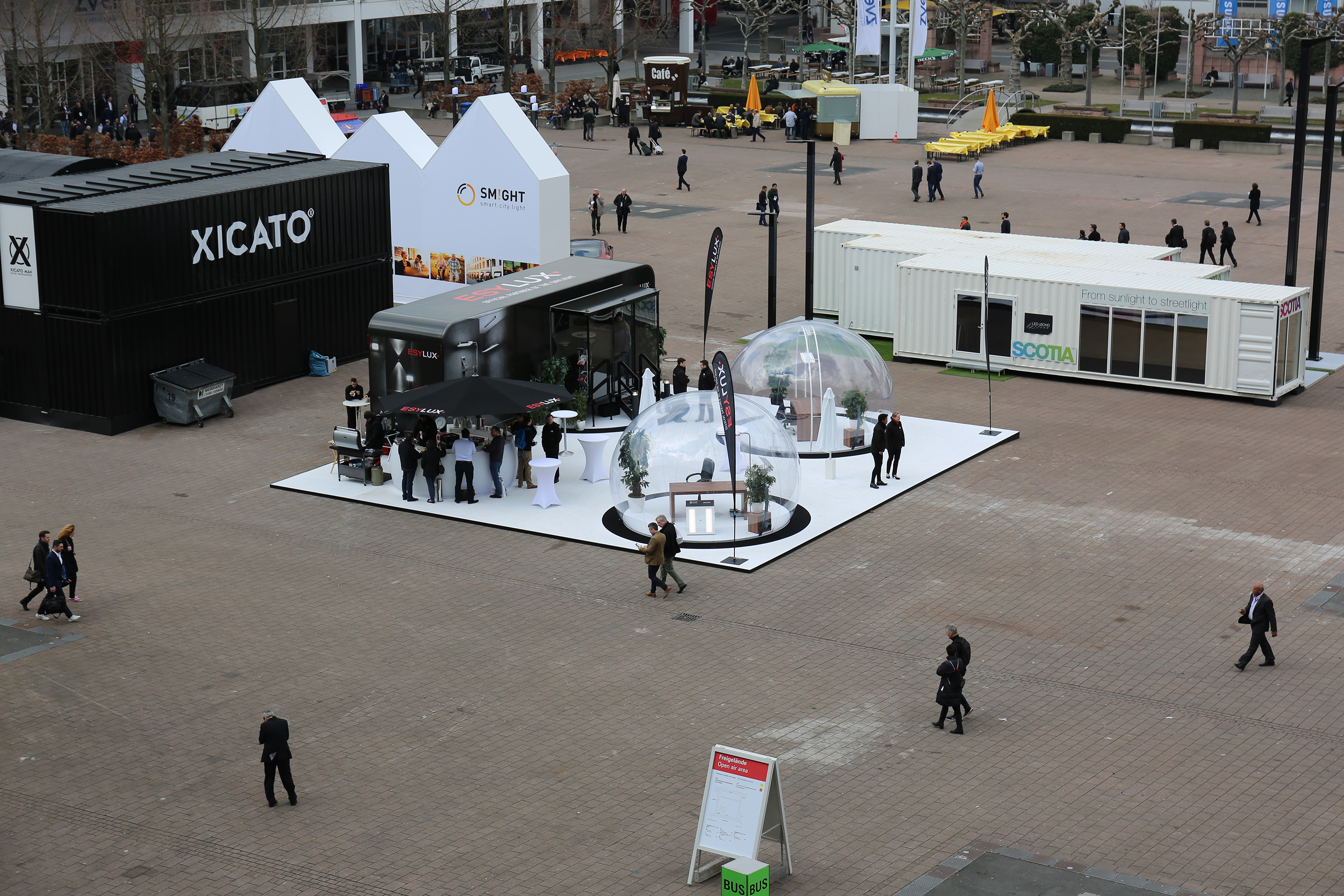 Messe-Außenplatz von oben: Im Fokus der Messestand von Esylux mit schwarzem Messeanhänger und zwei Glasiglus in denen Büromöbel ausgestellt werden.