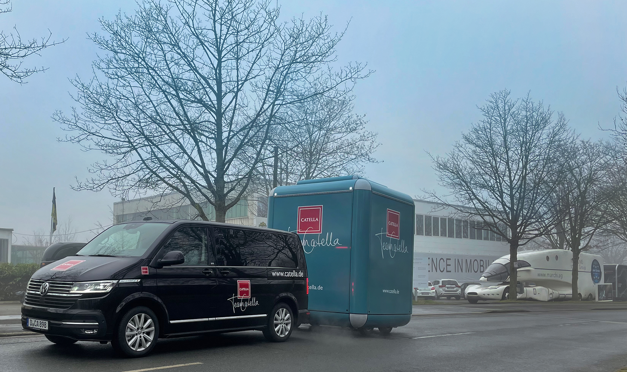 Schwarzer PKW zieht türkisen Promotion-Cube-Anhänger durch Winterlandschaft.