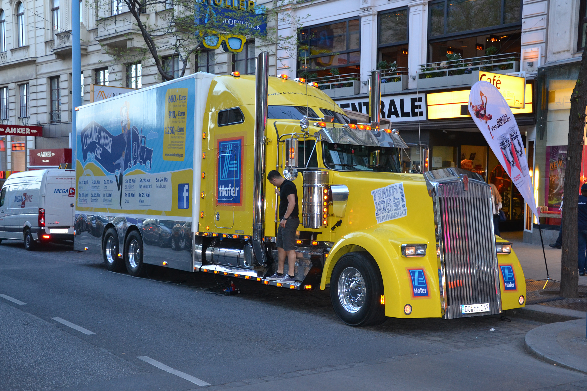 Gelber Showtruck mit Merchandise-Anhänger auf städtischer Straße mit Läden.