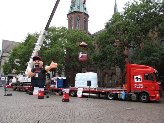 Stadtplatz: Kran hebt einen Pavillon von einem Laster. Auf dem Laster steht ebenfalls ein riesige Eisblock-Figur. Davor steht eine 5 Meter große aufgeblasene Moderator-Figur. 