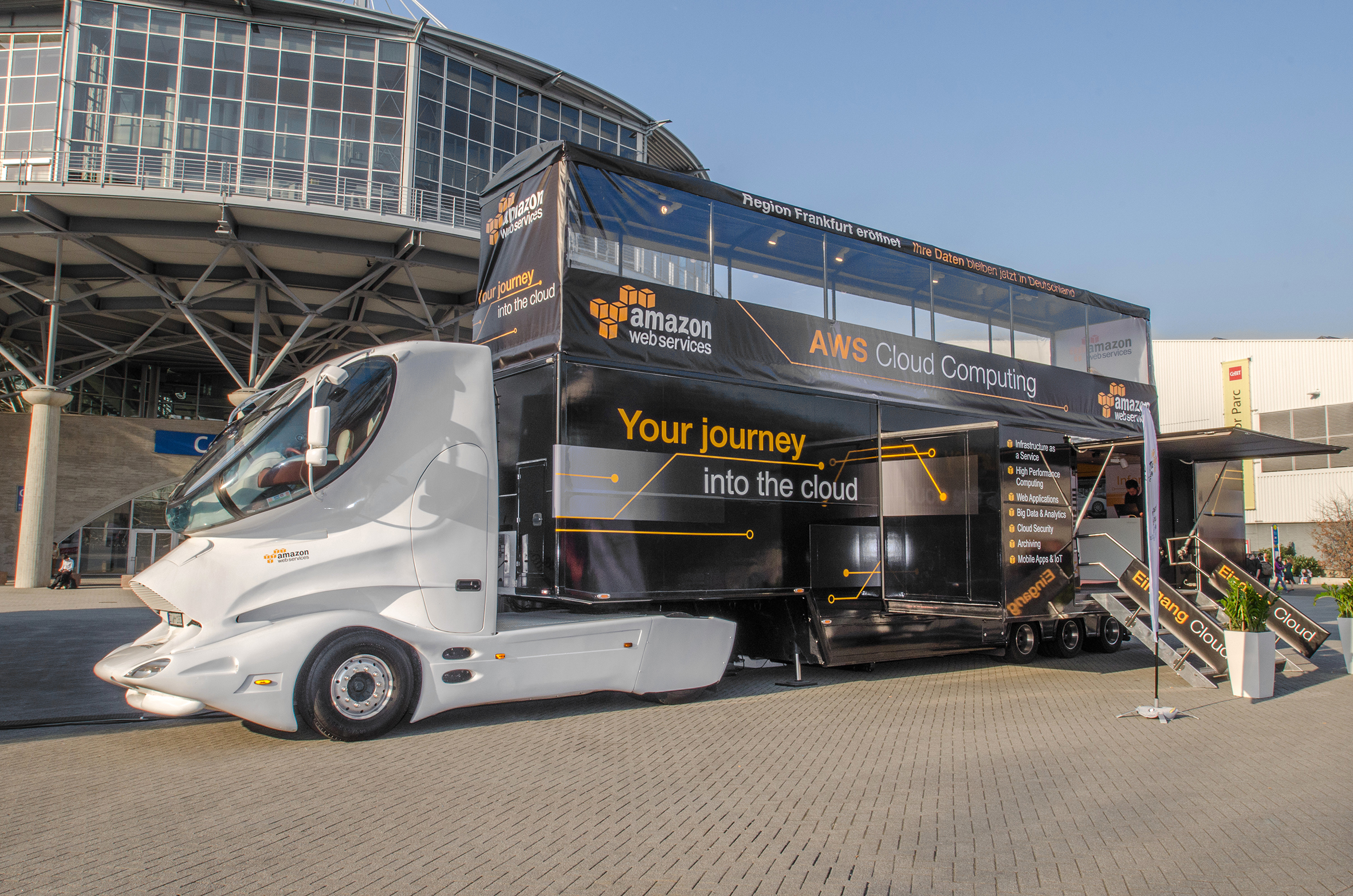 AWS Cloud Computing Showtruck mit weißem Design-Cockpit. Schwarzer Sattelauflieger mit orangenen Werbesprüchen und hochgefahrenen Außenwänden. 