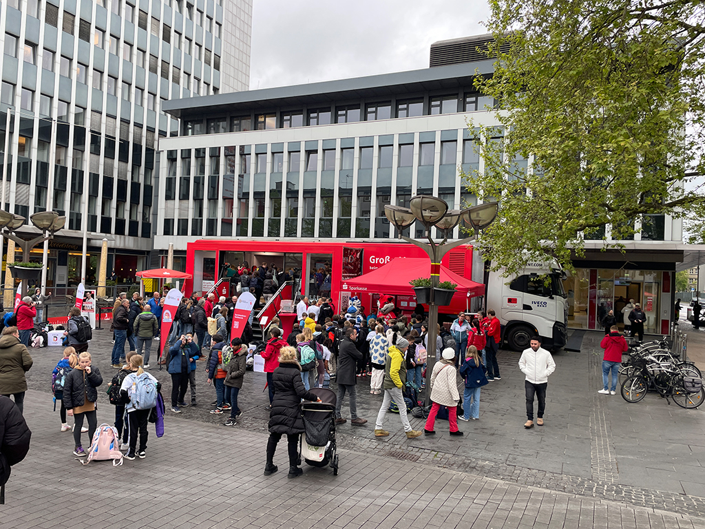 Roter Showtruck mit vielen Besuchern auf einem urbanen Platz vor modernen Gebäuden.