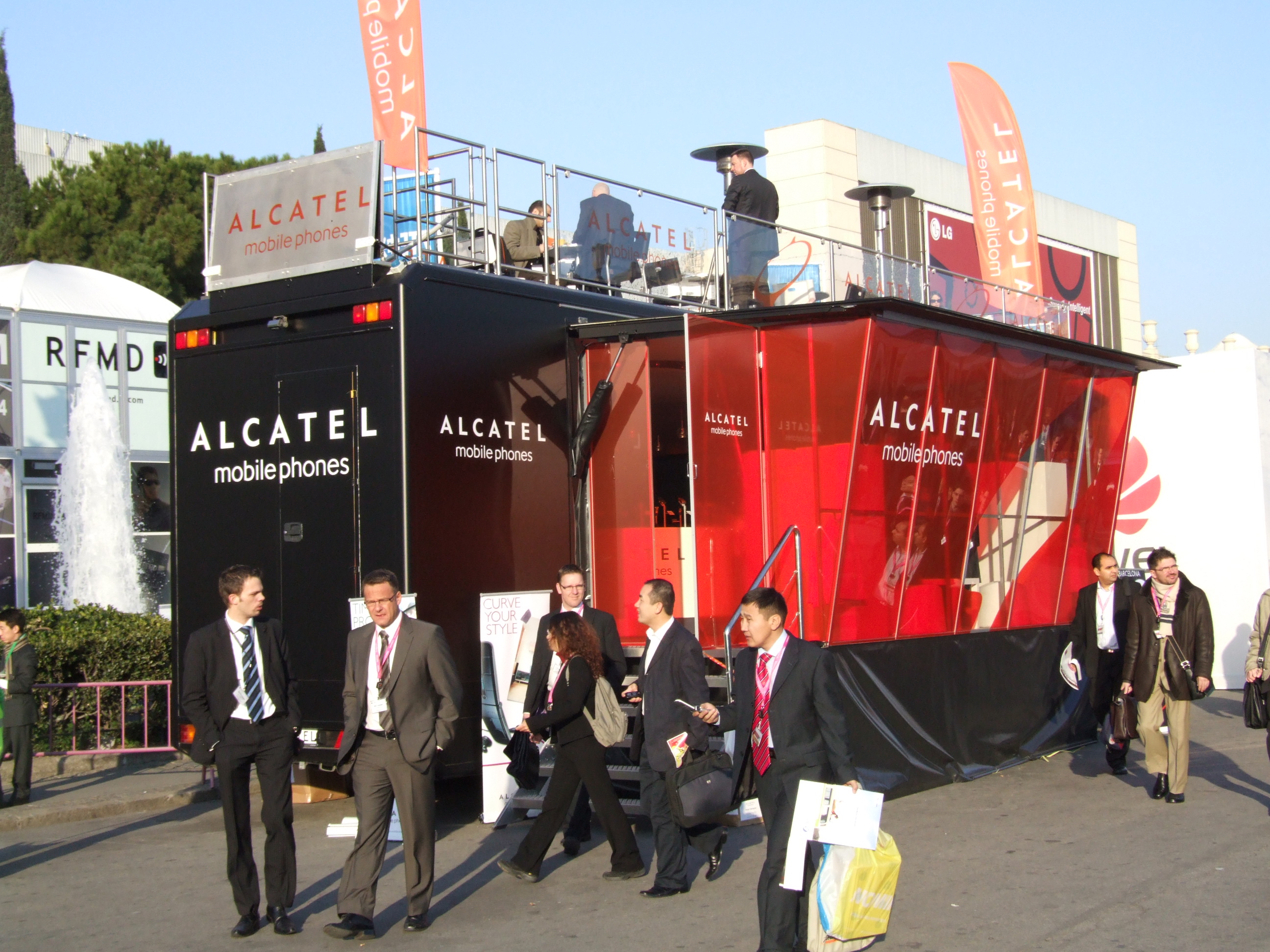 Alcatel Messetruck mit schwarz-rotem Branding und Dachterrasse umgeben von Besuchern. 