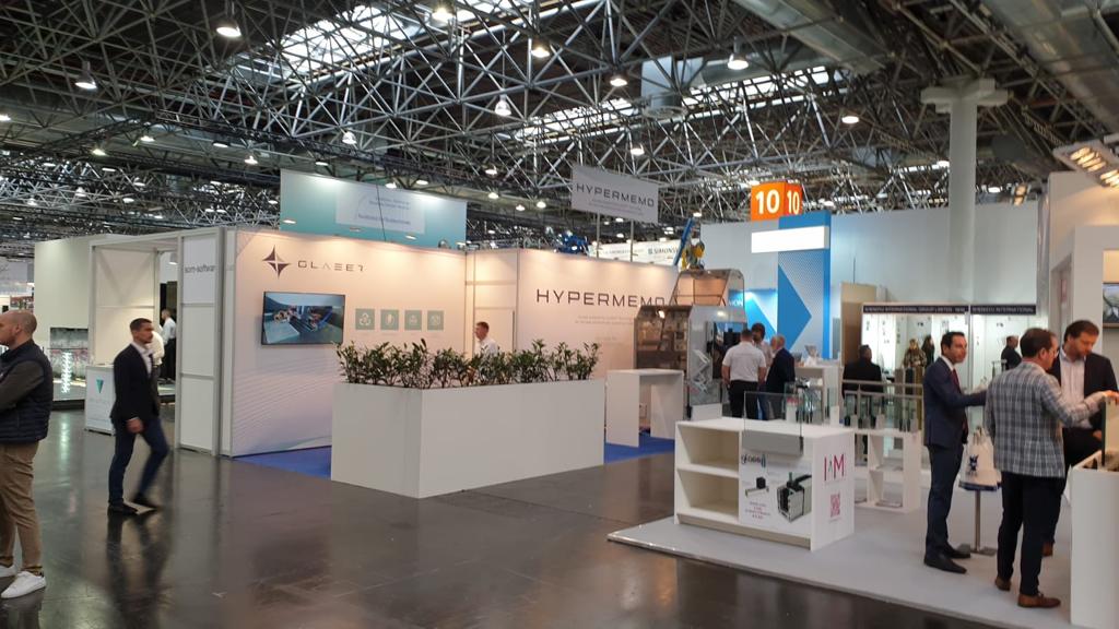 Moderner Messestand in Industriehalle: Blauer Boden, weiße gebrandete Trennwände, futuristische Tresen und Tische sowie Hochbeete.