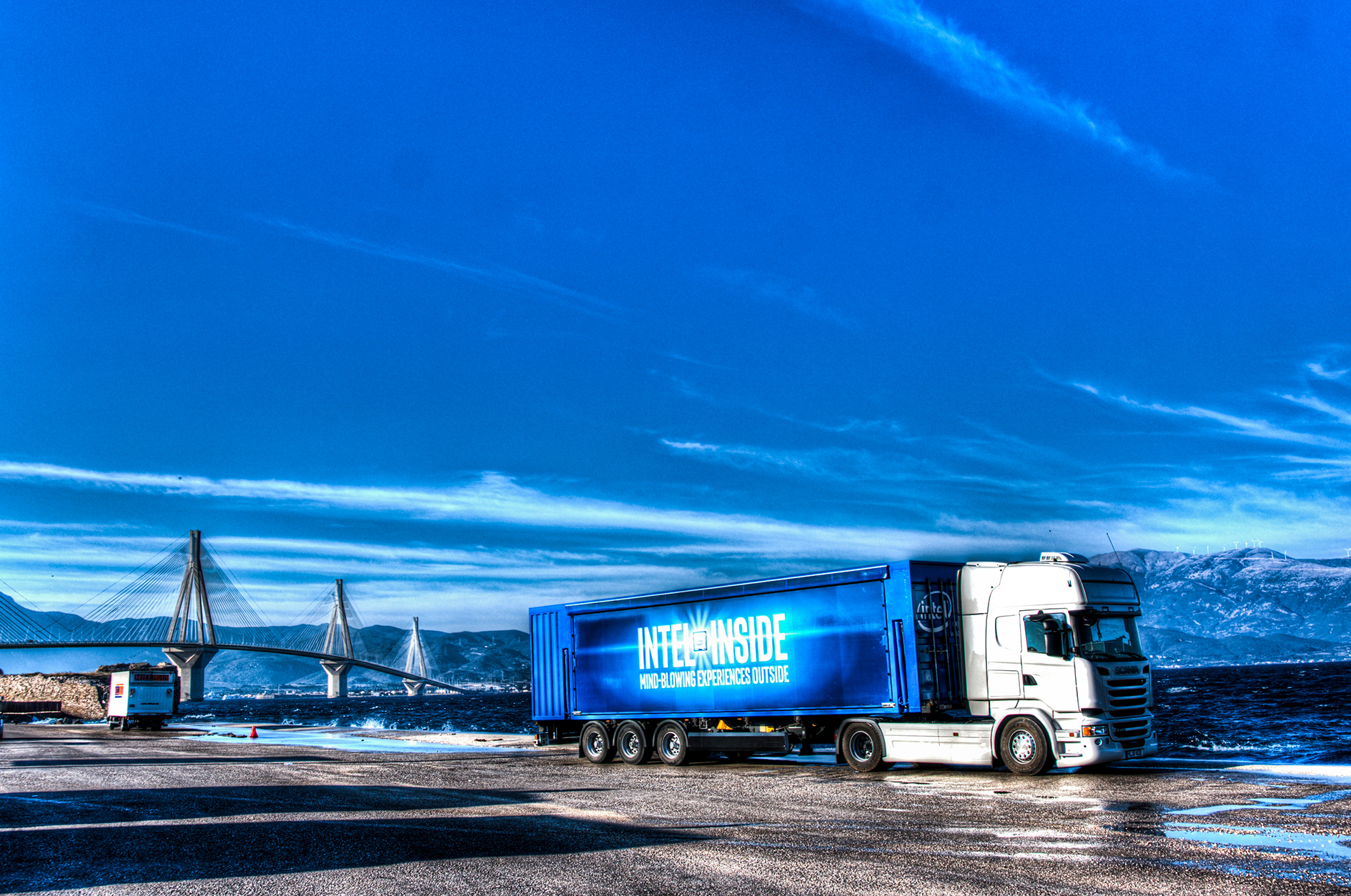 Showtruck mit blauem LED-Display an Küstenlandschaft vor moderner Brücke.