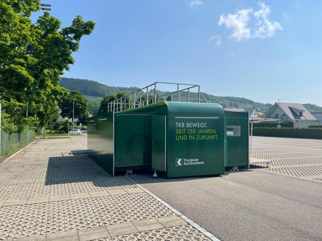 Grüner Showcontainer mit TKB-Branding und ausgefahrenen Seiten steht auf einem Parkplatz.