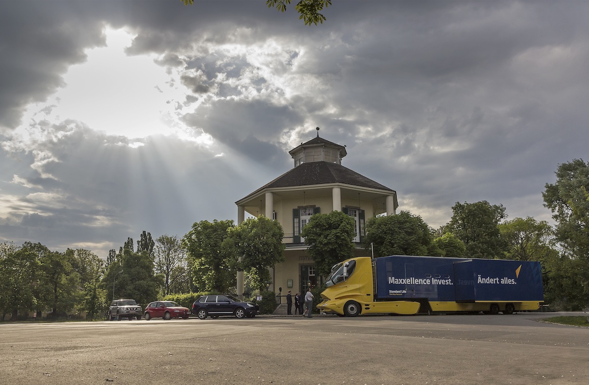 Gelber Showtruck mit dunkelblauem Auflieger vor historischem Rundbau unter dramatischem Himmel.