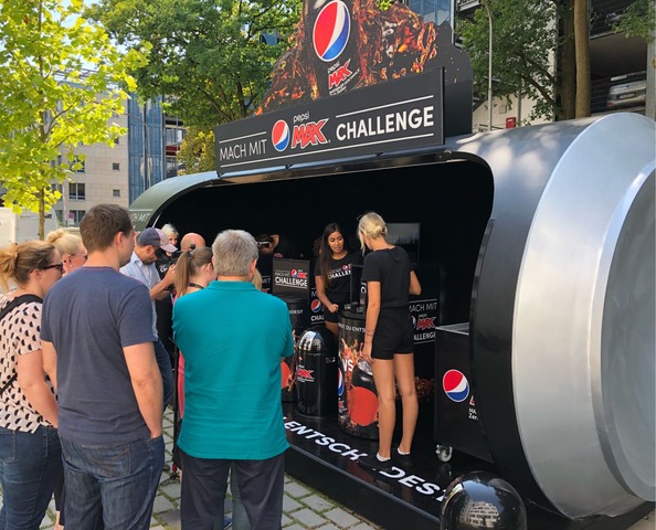 Begehbarer PKW-Anhänger in Pepsi-Form: Promoterinnen verteilen Pepsi-Dosen an Passanten.