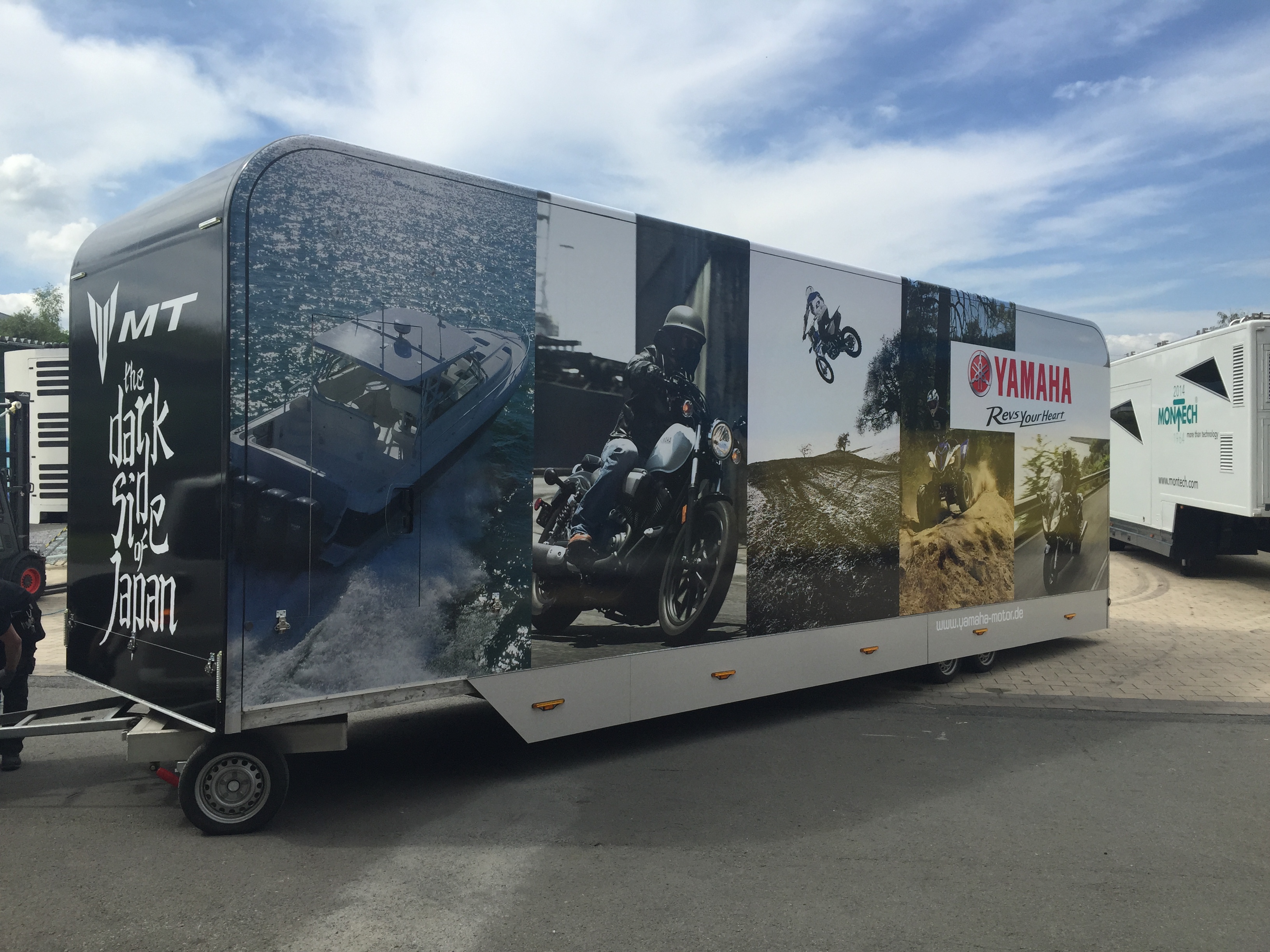 Großer schwarzer Werbeanhänger mit Motorrädern, Booten und Yamaha-Logo auf Parkplatz.