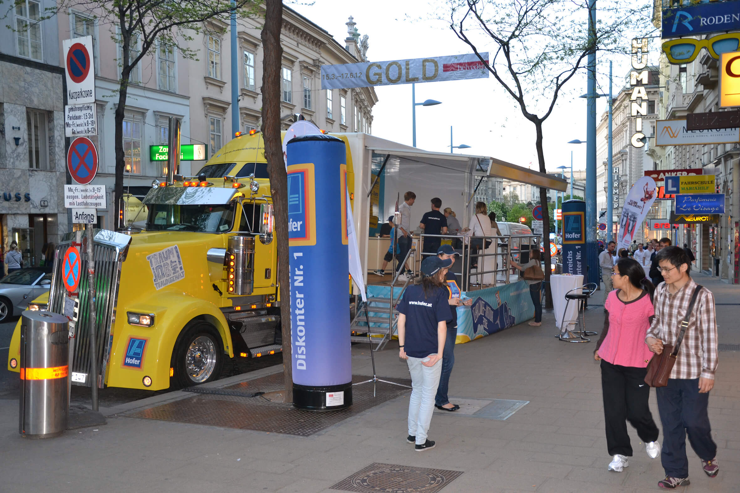 Gelber US-Truck mit offenem Bühnenauflieger als Aldi-Recruiting-Roadshow auf einer Wiener Einkaufsstraße, mit Promotern und Infopylonen davor.