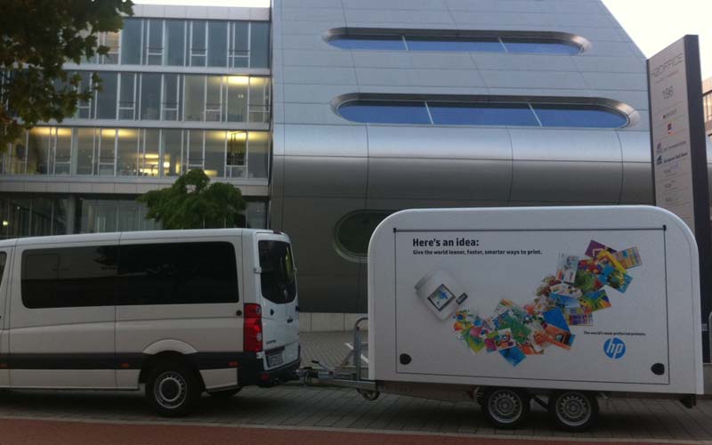 Weißer HP-Promotionanhänger mit Druckmotiven, gezogen von einem Kleinbus vor einem modernen Bürogebäude.