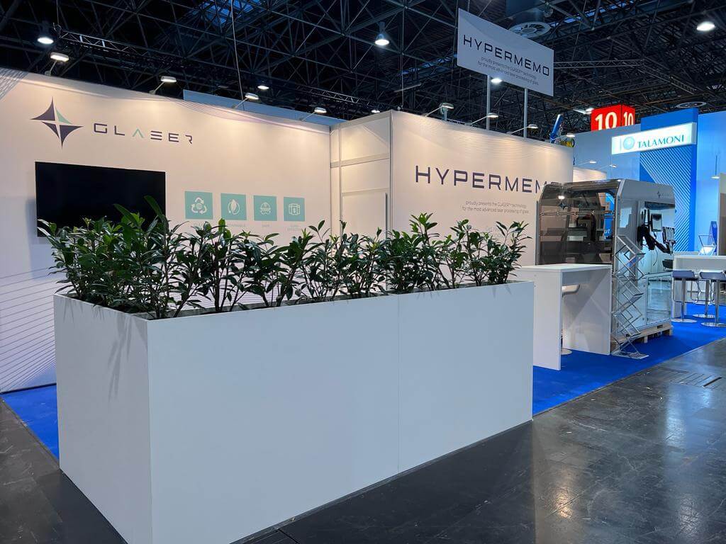 Messestand von Glaser mit weißen Wänden, Pflanzkübel und ausgestellter Lasermaschine in einer Messehalle.