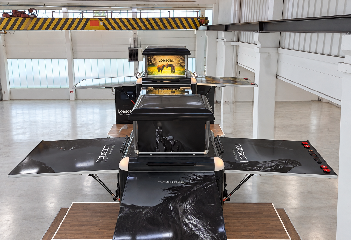 Two black Marchi Cube sales units for Loesdau, with horse-themed graphics and fold-out side counters, set up on wooden platforms in a bright industrial hall.