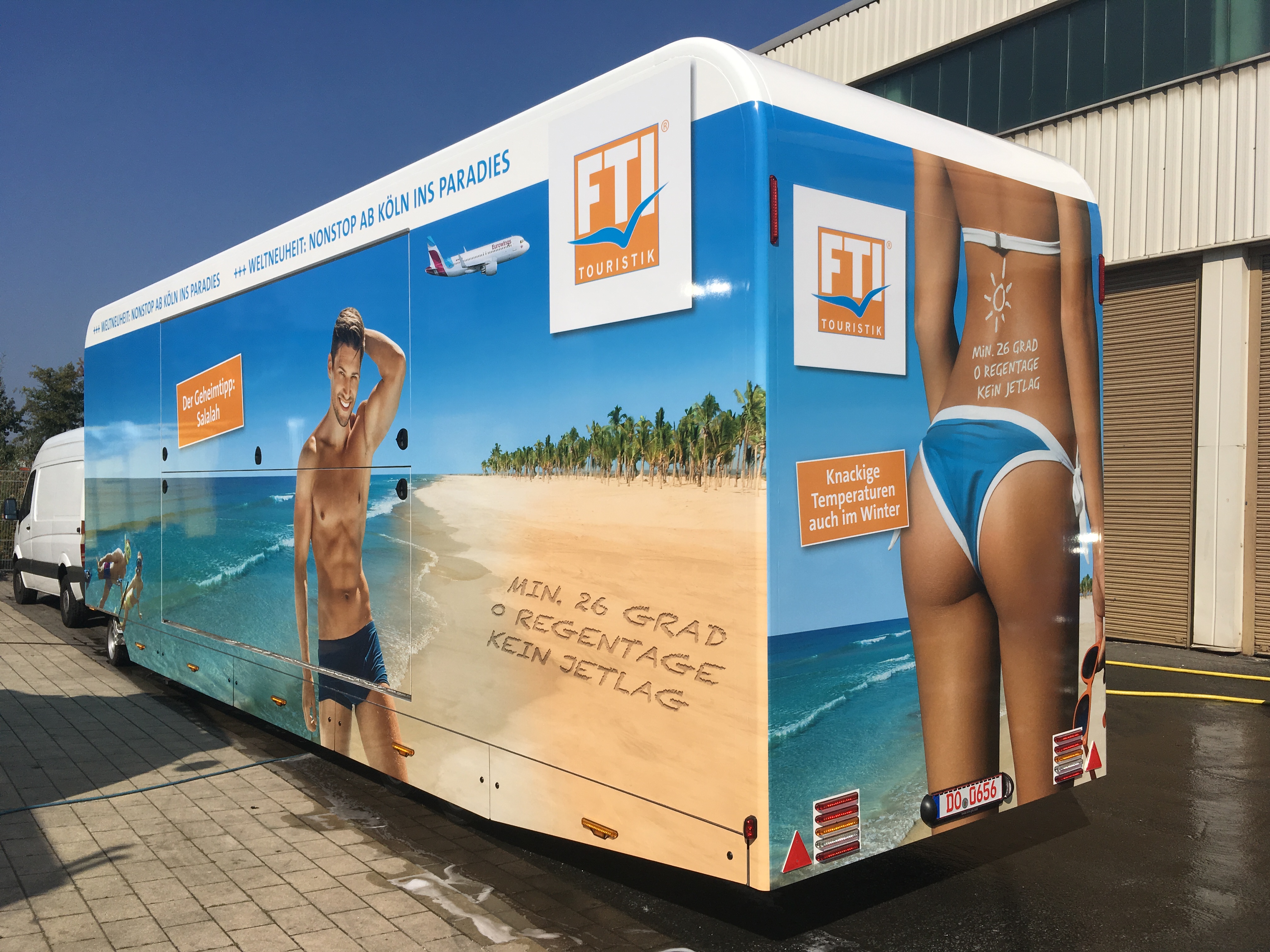 FTI Touristik trailer with a beach and palm tree wrap, towed by a white van and parked beside an industrial building.