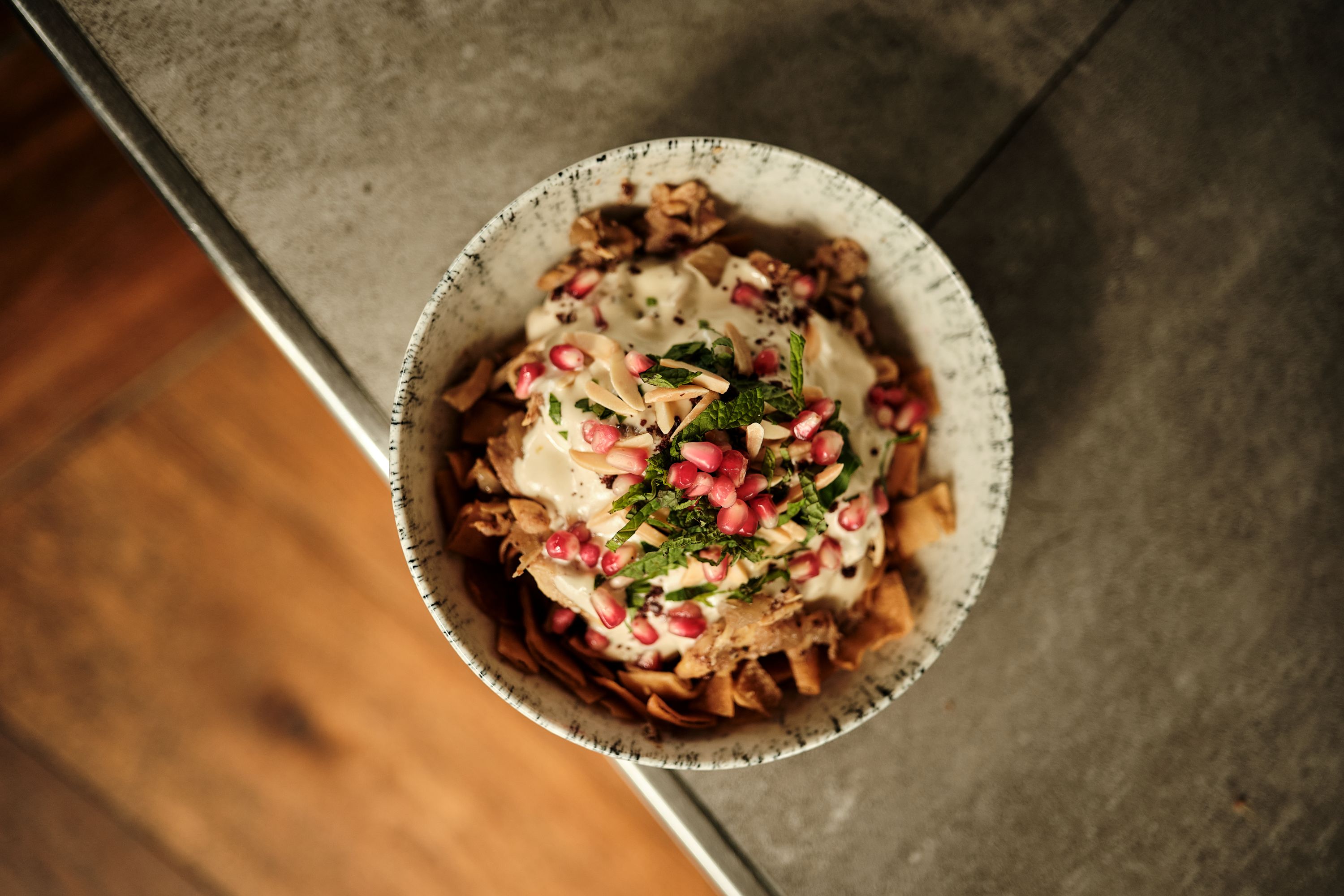 Bowl of food topped with white sauce, pomegranate seeds, chopped herbs, and toasted nuts on a gray surface.