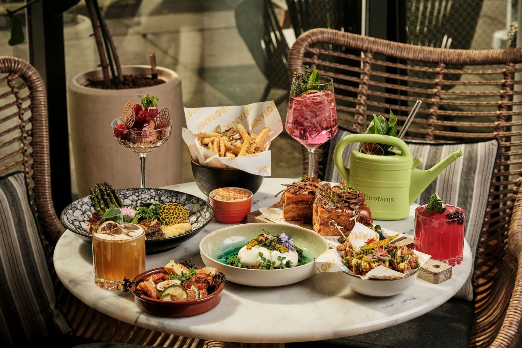 Table set with gourmet dishes and pink cocktails in a cozy rattan chair setting.
