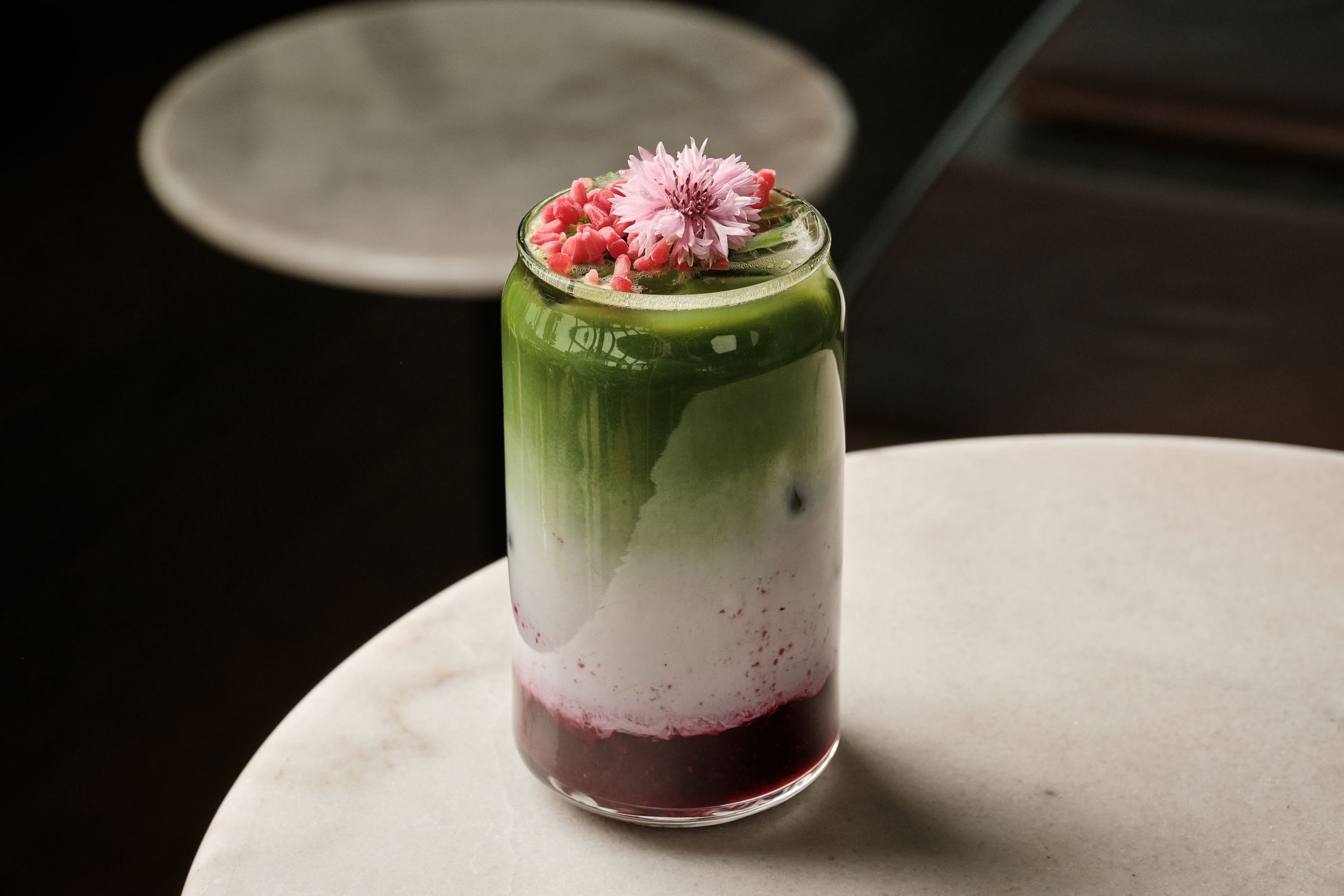 Layered iced matcha drink with milk and berry syrup, topped with pink flower and red buds in a clear glass.