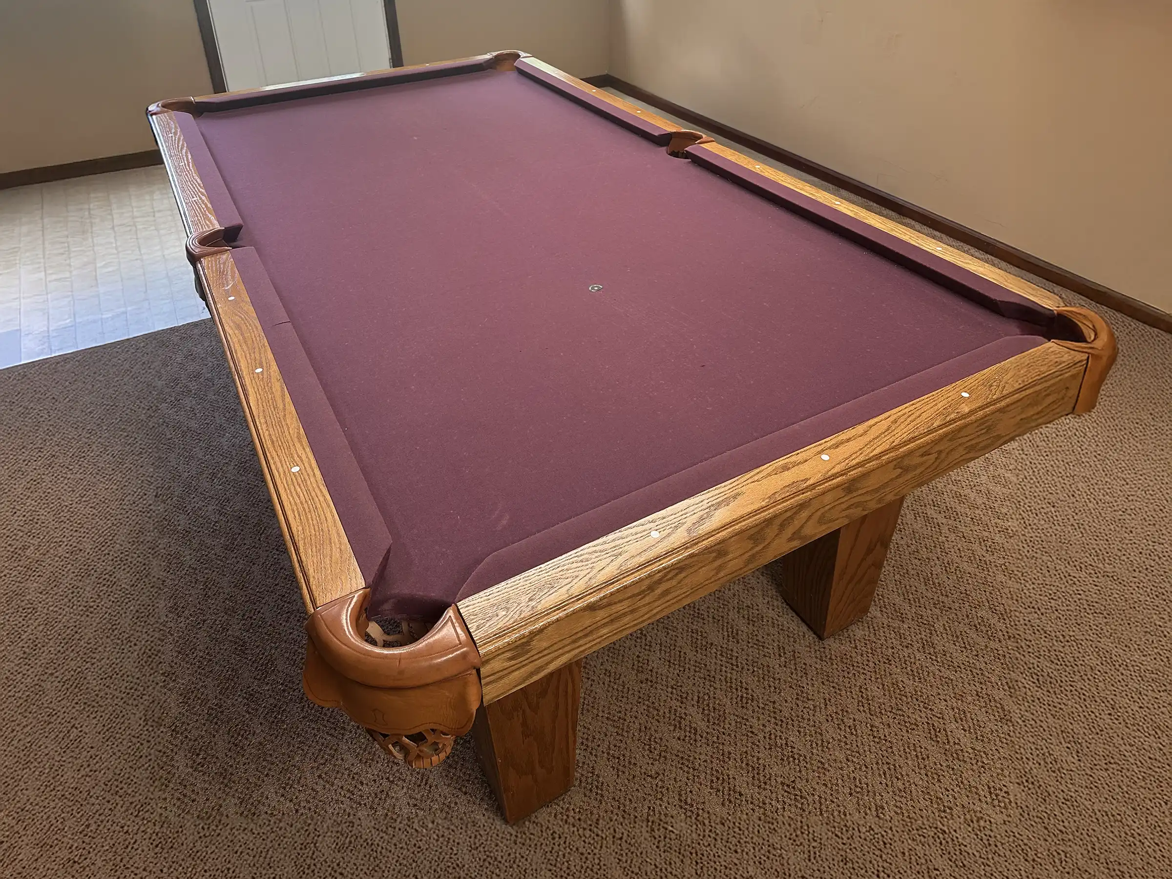 Olhausen Sheraton 8-foot pool table with maroon red felt, oak frame, leather pockets, and sturdy wooden legs, placed in a carpeted game room.