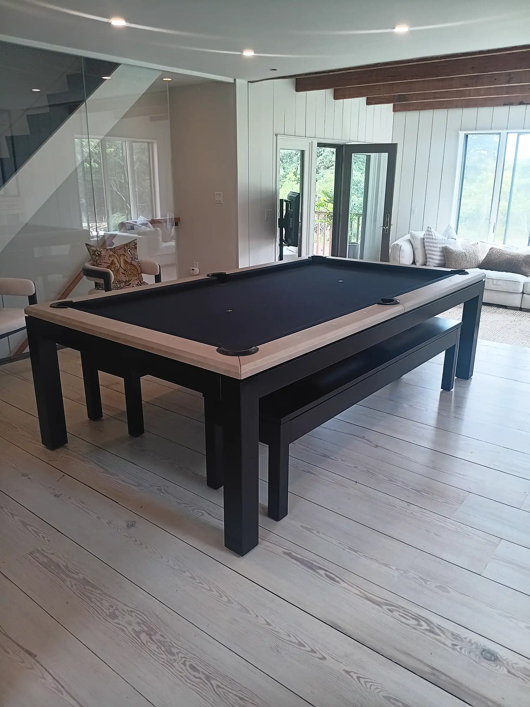 Pool table with black felt and light wood rails, featuring a modern black frame and a matching bench in a minimalist room with wood floors.