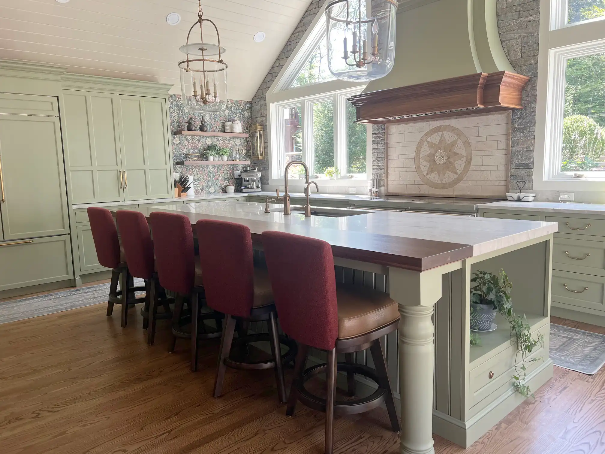 Spacious kitchen with sage green cabinets, a large island with a wood countertop, six red upholstered stools, and large windows providing natural light.