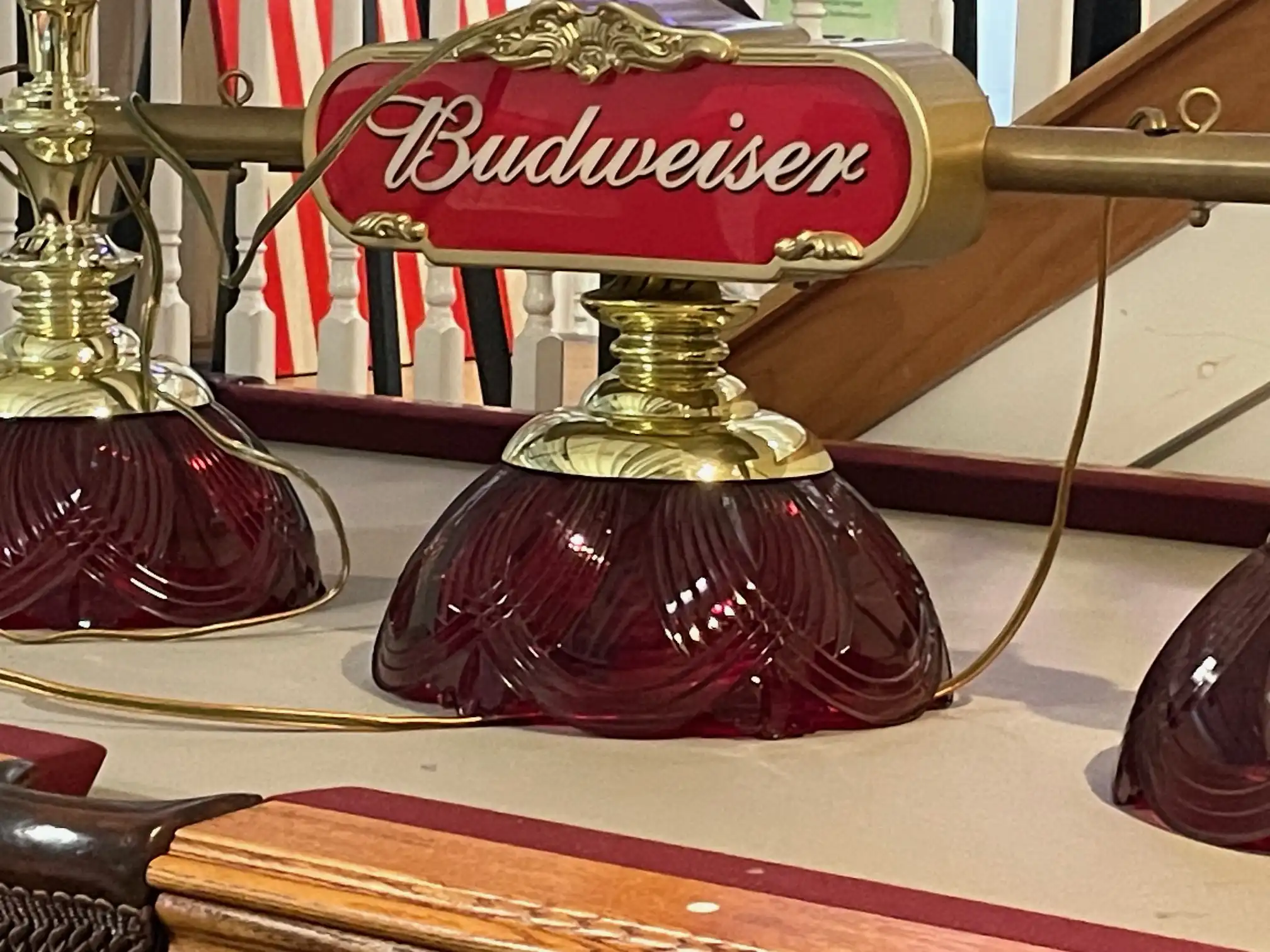 Vintage Budweiser pool table light with gold hardware and deep red glass shades hanging over tan felt, set against staircase and railing backdrop.