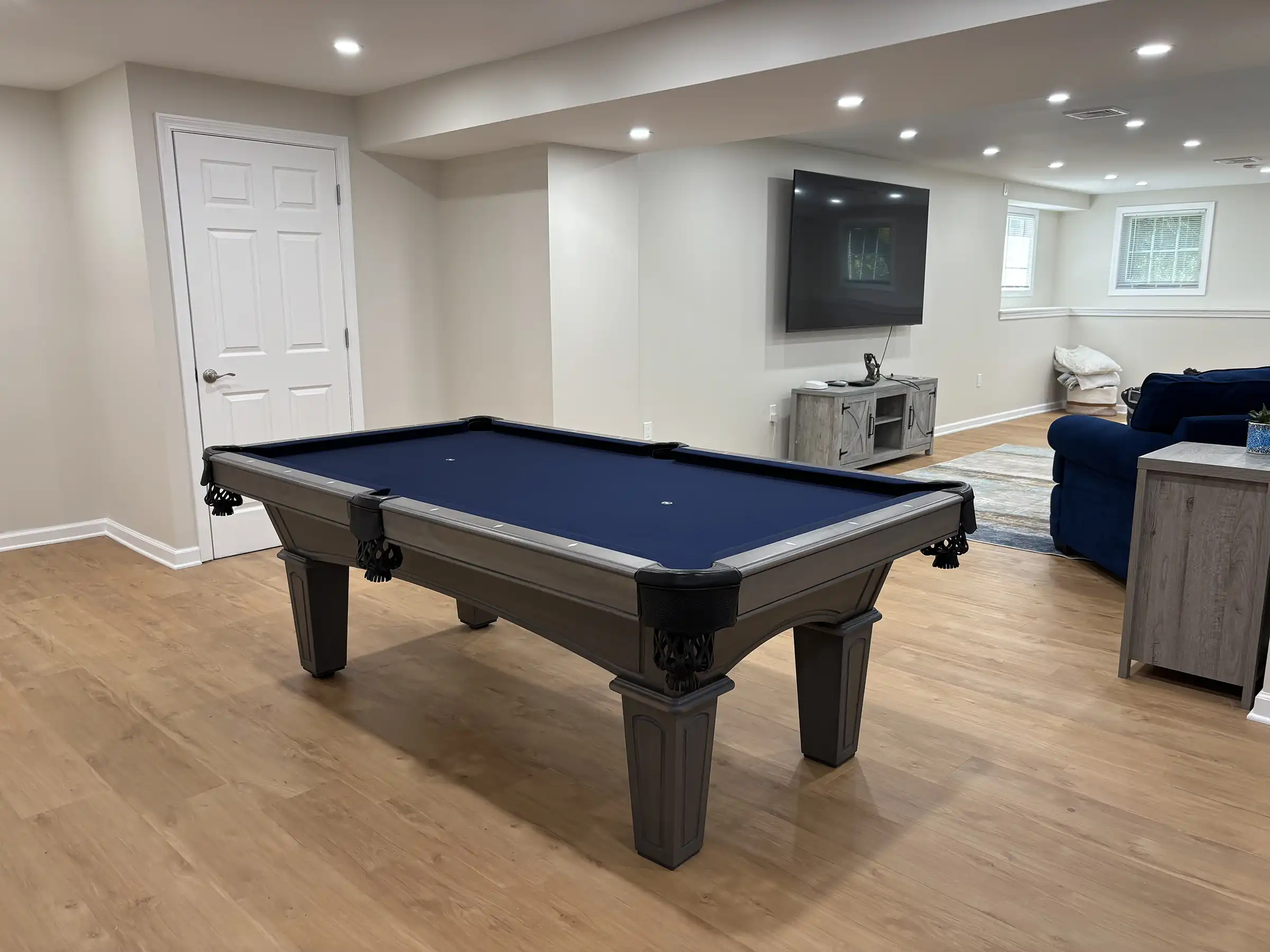 A pool table with a dark blue felt surface and a gray wooden frame sits in a bright, modern basement with light wood flooring.