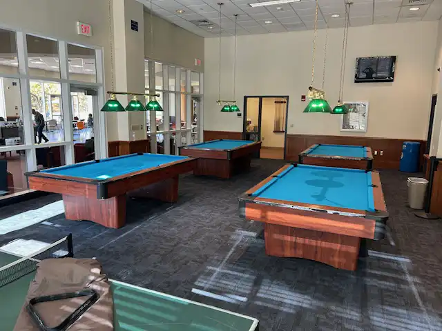 Four billiards tables with blue felt and green overhead lights in a spacious, well-lit game room with large windows and glass doors in the background.