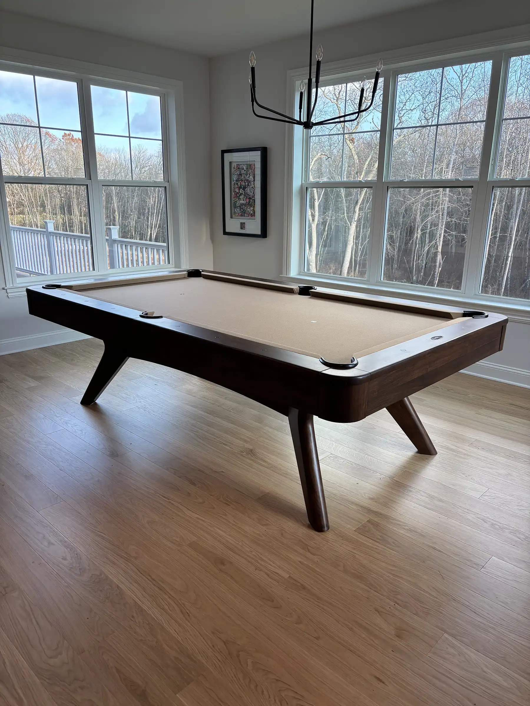 Mid-century modern pool table with dark wood finish and tan (light brown) felt in a bright room with large windows overlooking trees.