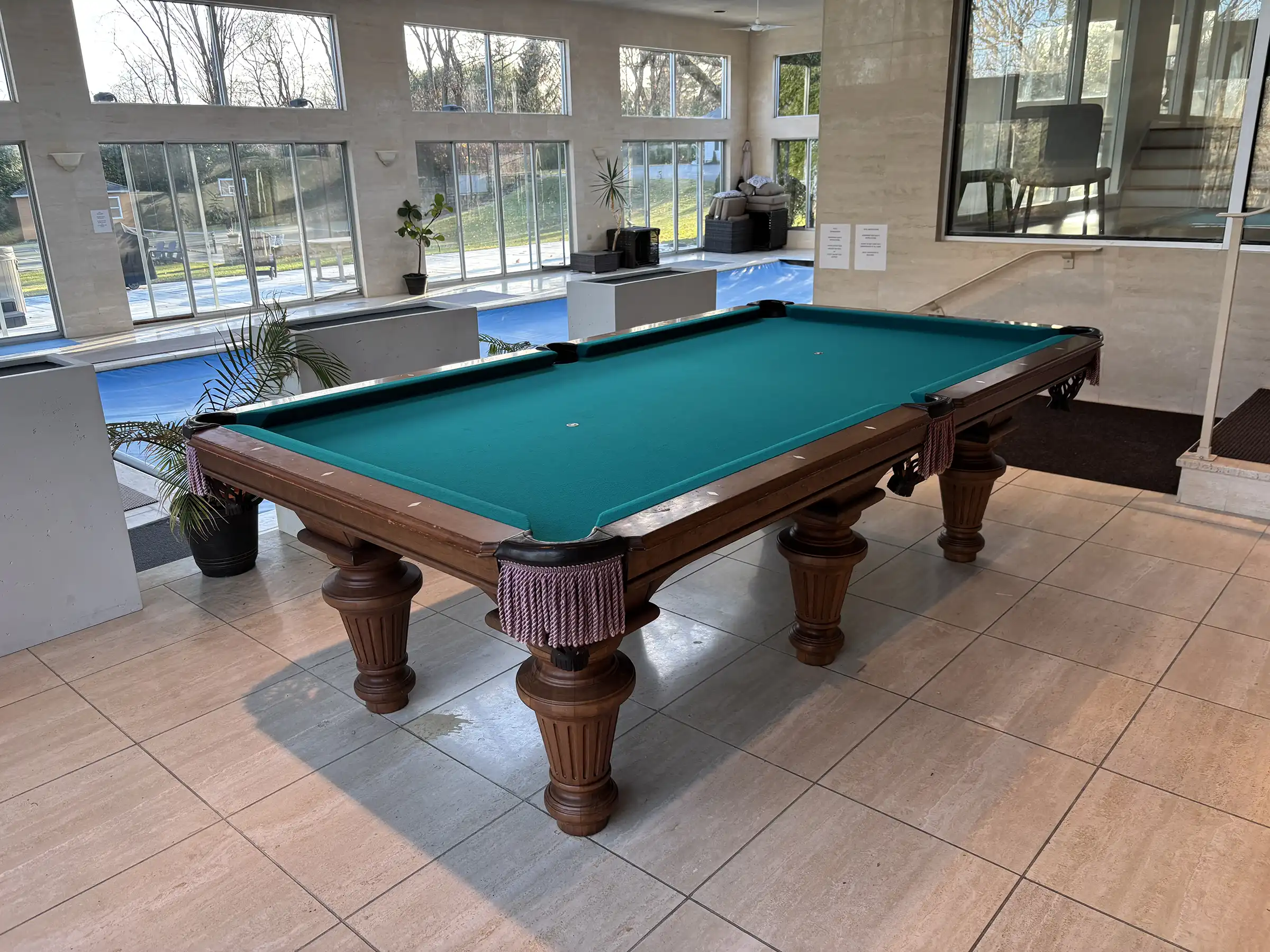 Traditional pool table with ornate carved legs and green felt, set in a bright luxury indoor swimming pool room with large windows.