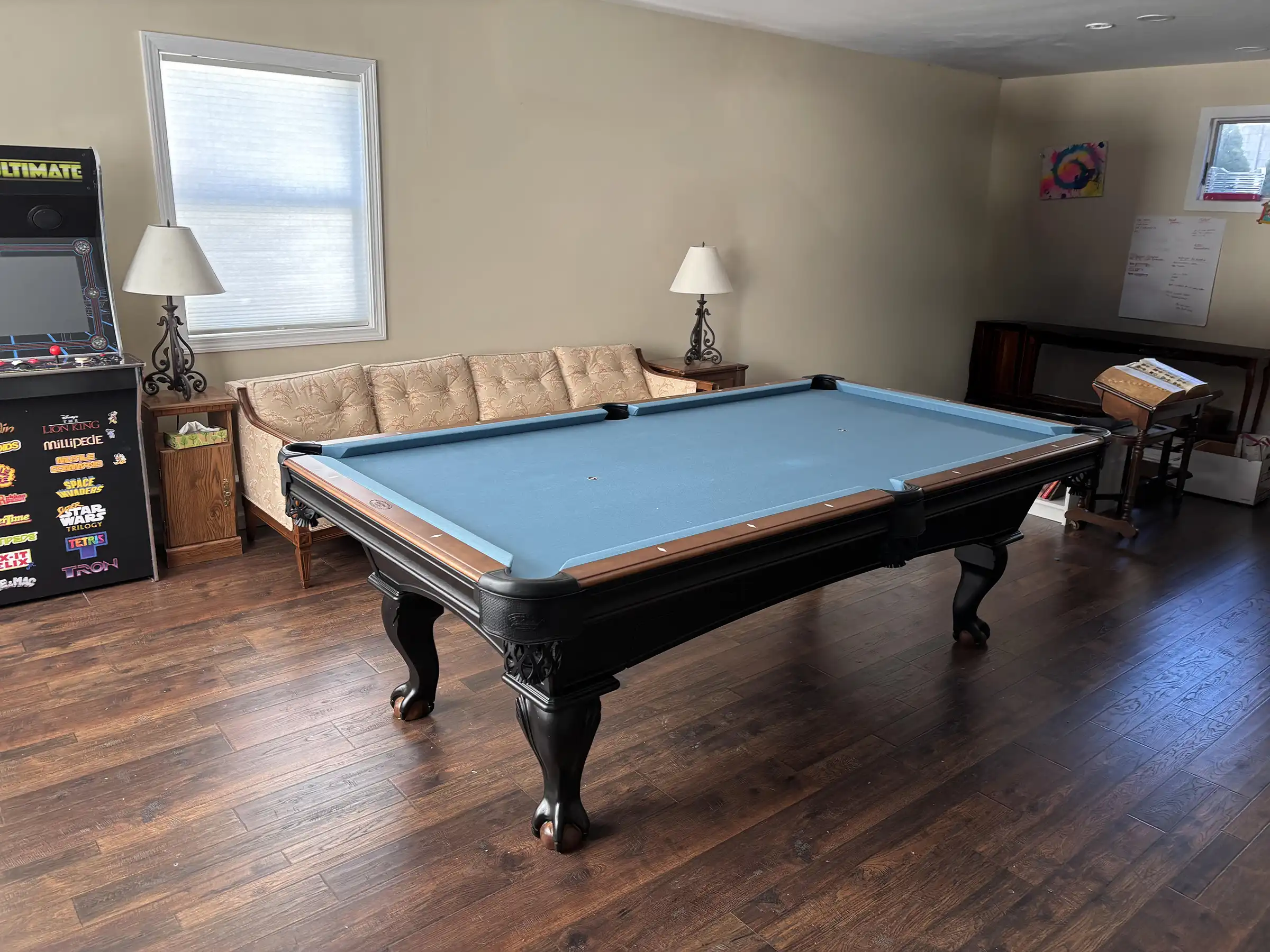 Classic black pool table featuring light blue felt and carved legs, near a vintage sofa and an arcade machine in a bright home game room.