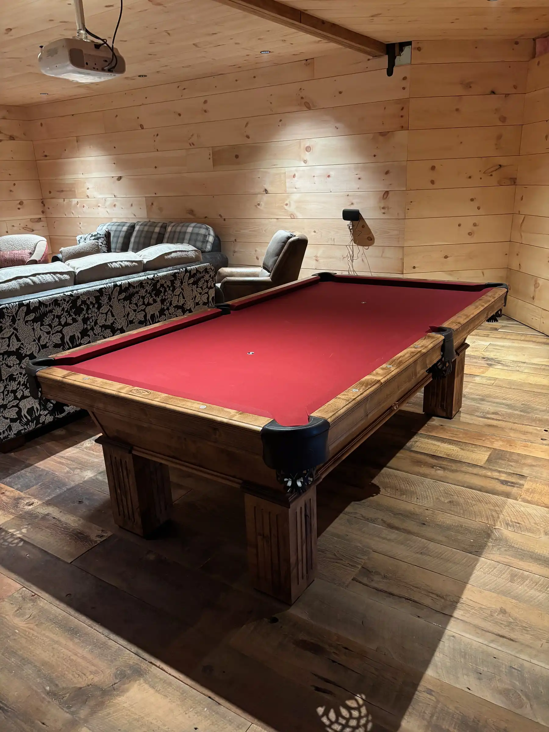 Rustic wooden pool table with red felt stands in a wood-paneled game room with plank floors, a patterned sofa, and a ceiling projector.